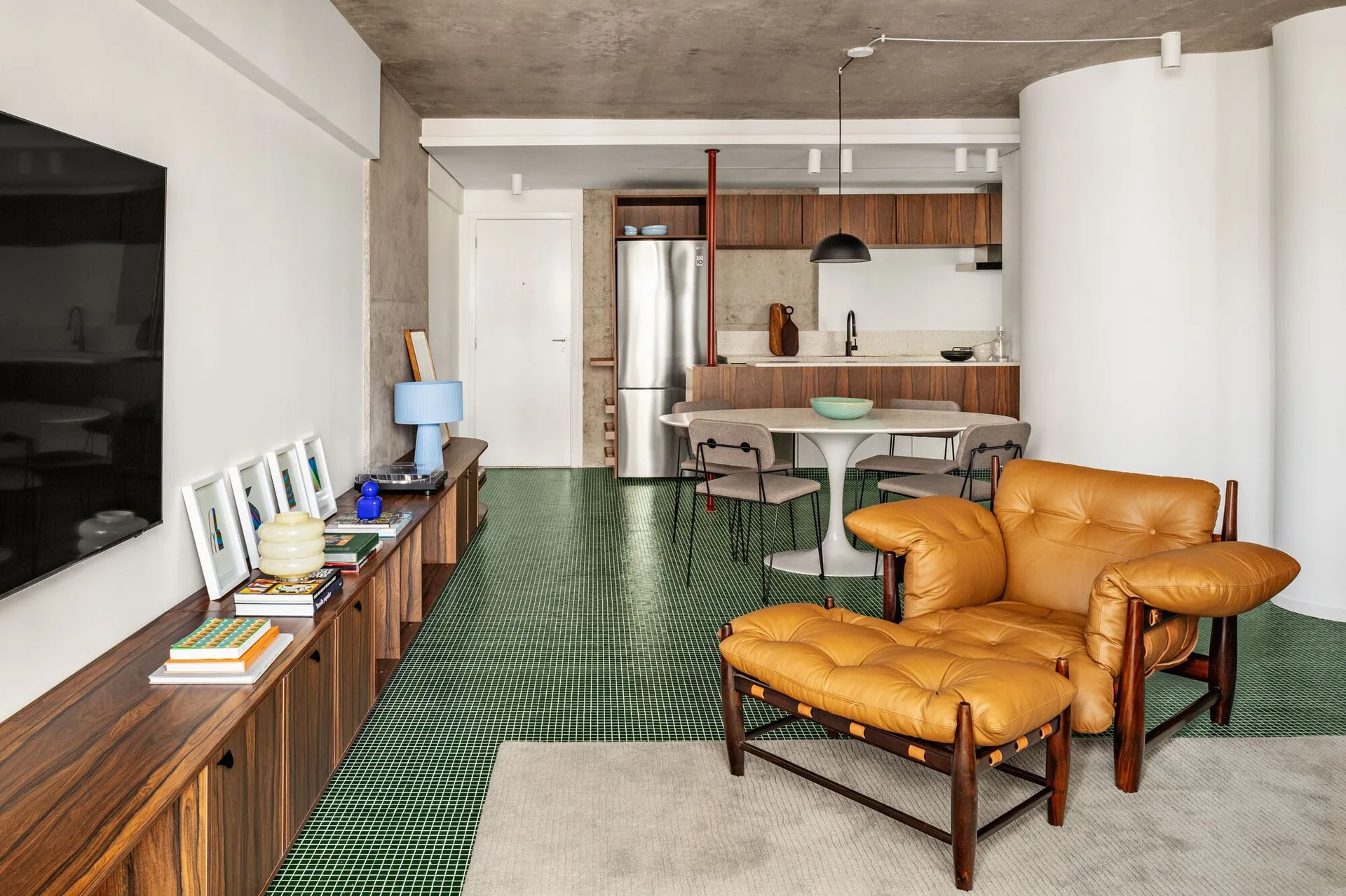 An open-concept living area features green tiled flooring, an exposed concrete ceiling, a tan leather armchair, and a wood media console.