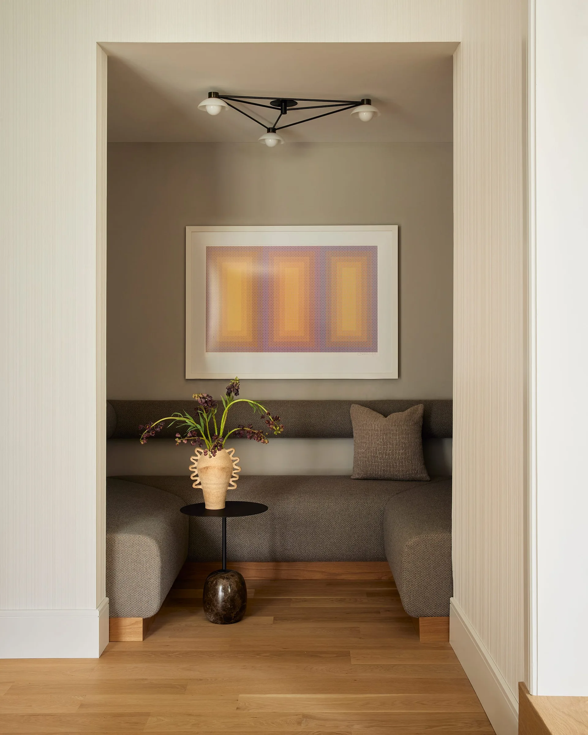 A recessed alcove features a U-shaped upholstered banquette, a small marble-base pedestal table, a modern ceiling light, and framed geometric artwork.