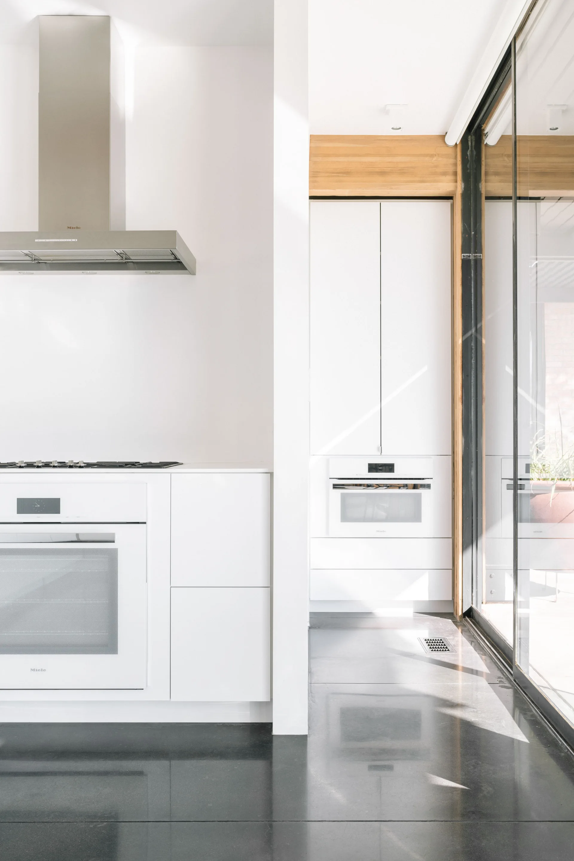 A minimalist white kitchen featuring flat-panel cabinets, built-in white ovens, and a polished dark grey floor next to a glass sliding door.