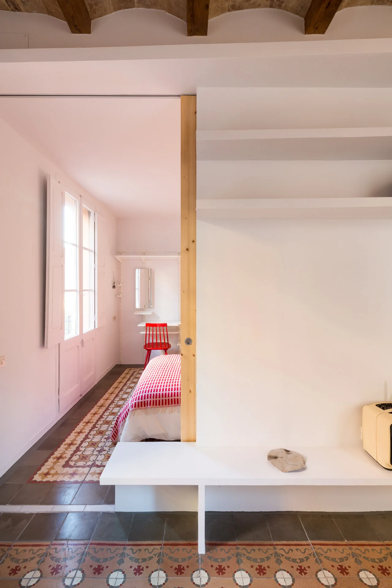 A minimalist bedroom viewed through a white partition, showing a bed with red patterned bedding, geometric tile floors, and exposed wooden beams.