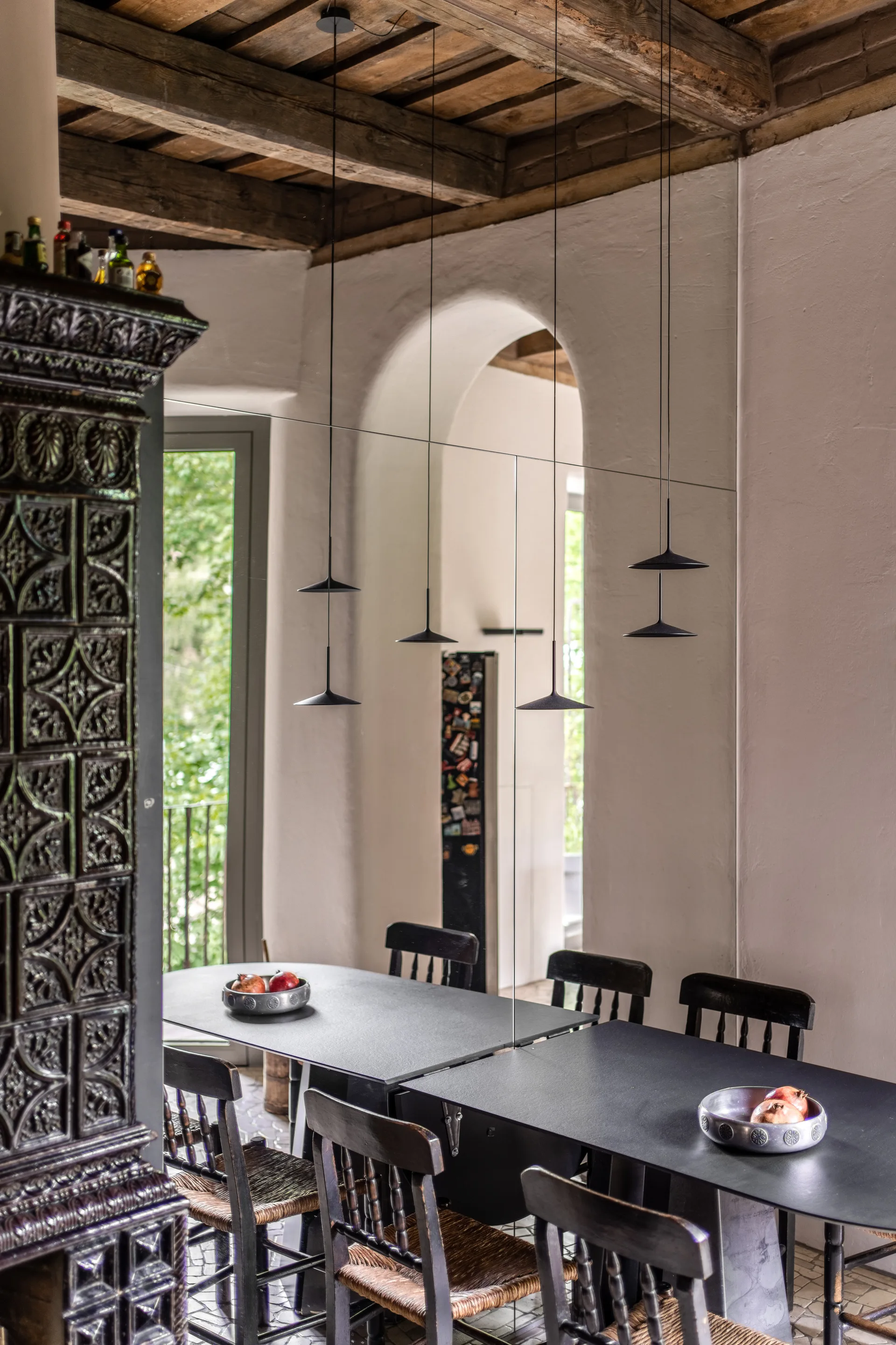 A dining room features a long black table, wooden chairs with woven seats, a large mirror, and slim black pendant lights under exposed ceiling beams.