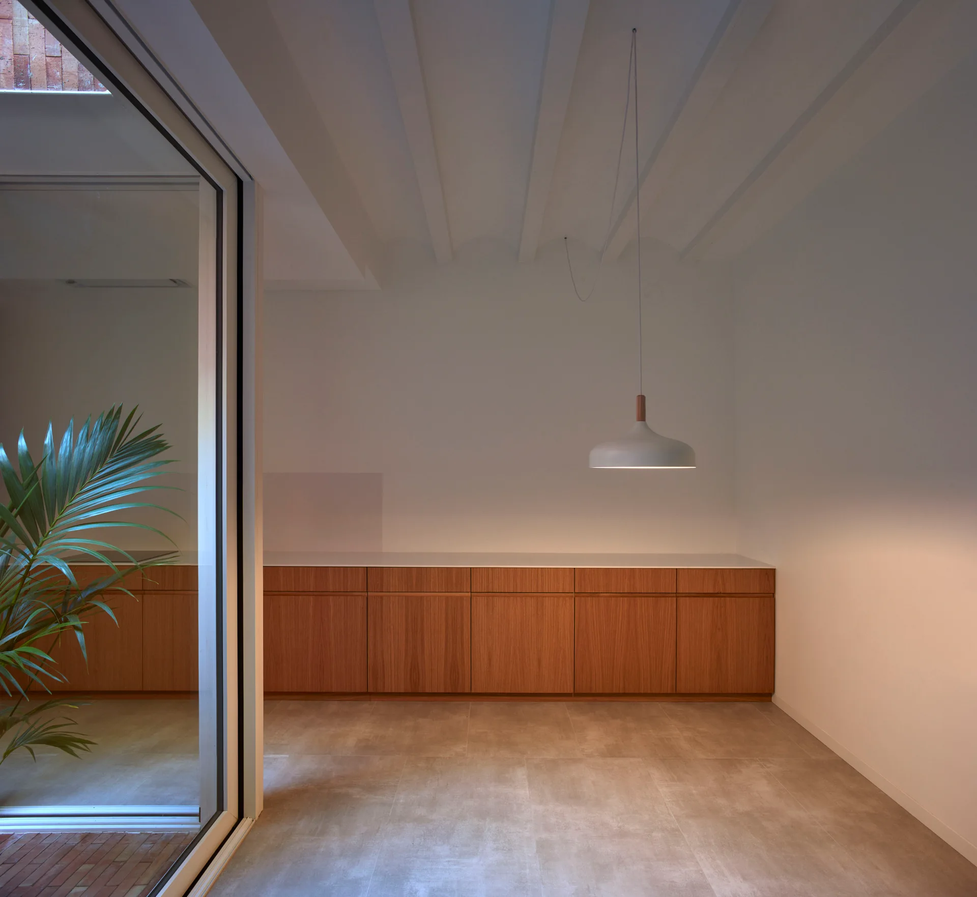 A minimalist kitchen featuring warm wood base cabinets, a white dome pendant light, white walls, and grey stone-like floor tiles.