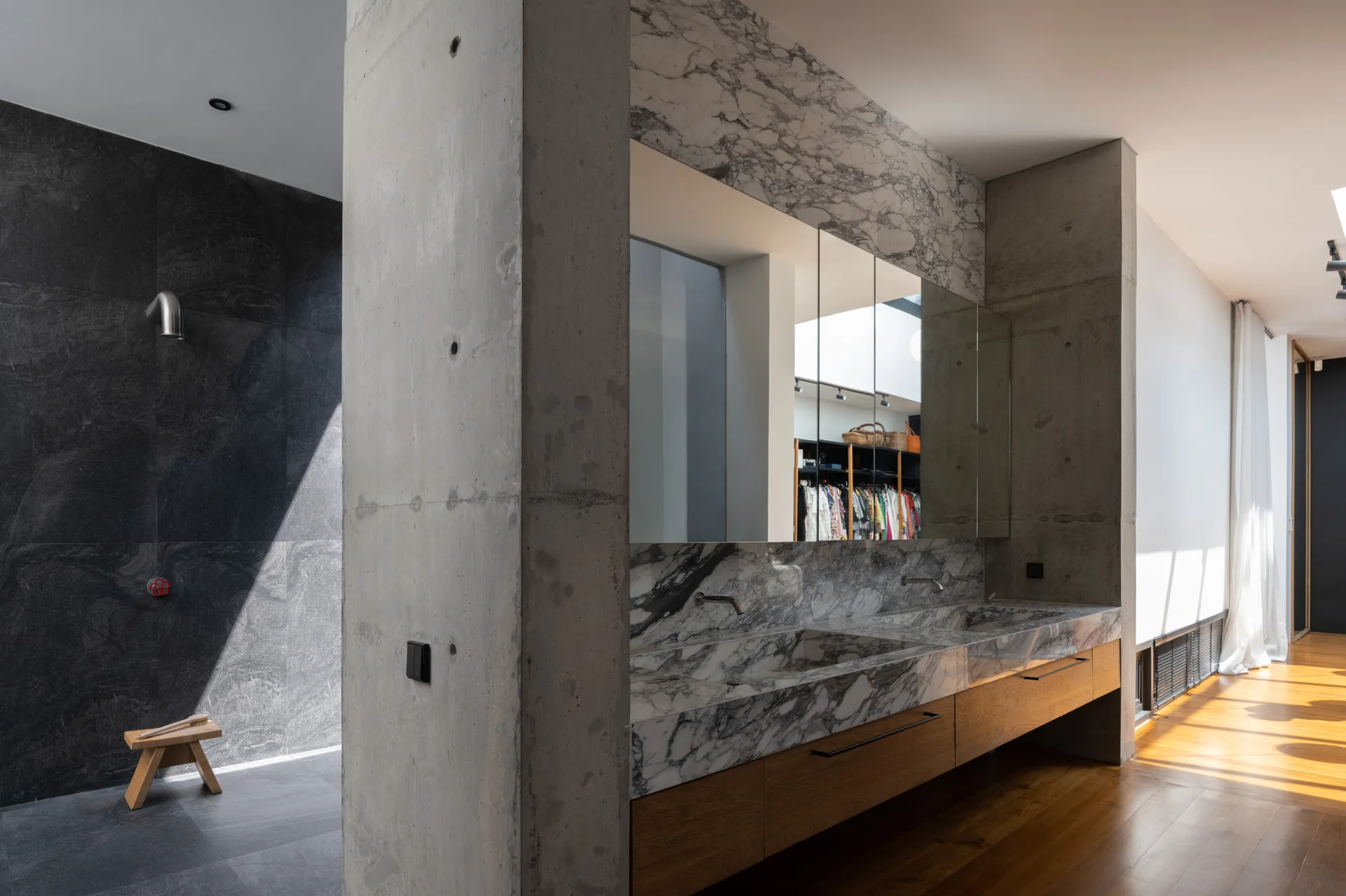 A modern bathroom featuring a heavily veined marble double vanity with wood cabinets set against raw concrete walls next to a dark slate shower.