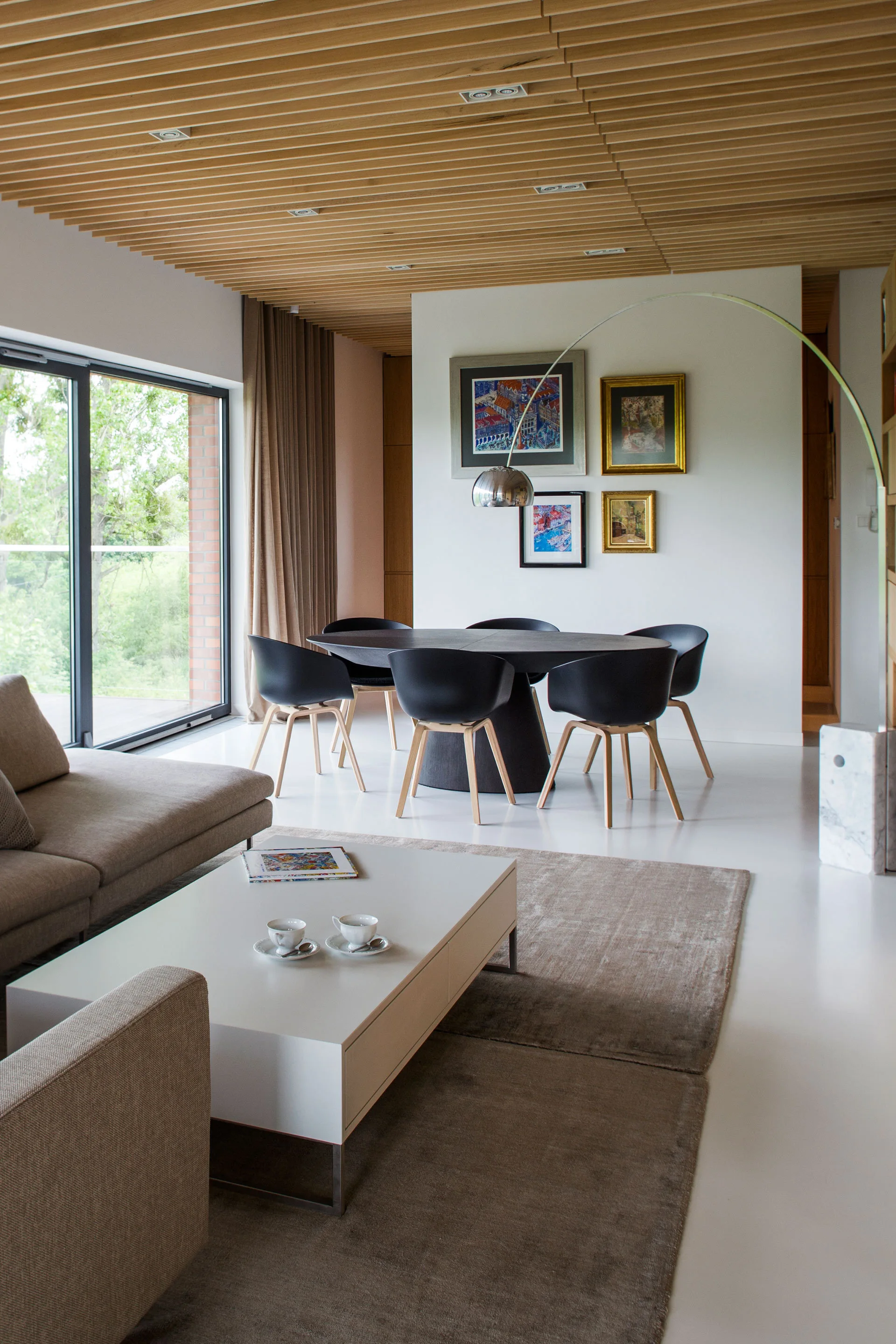 An open-plan dining and living space features a slatted wood ceiling, a round black dining table, and a grey sectional sofa.