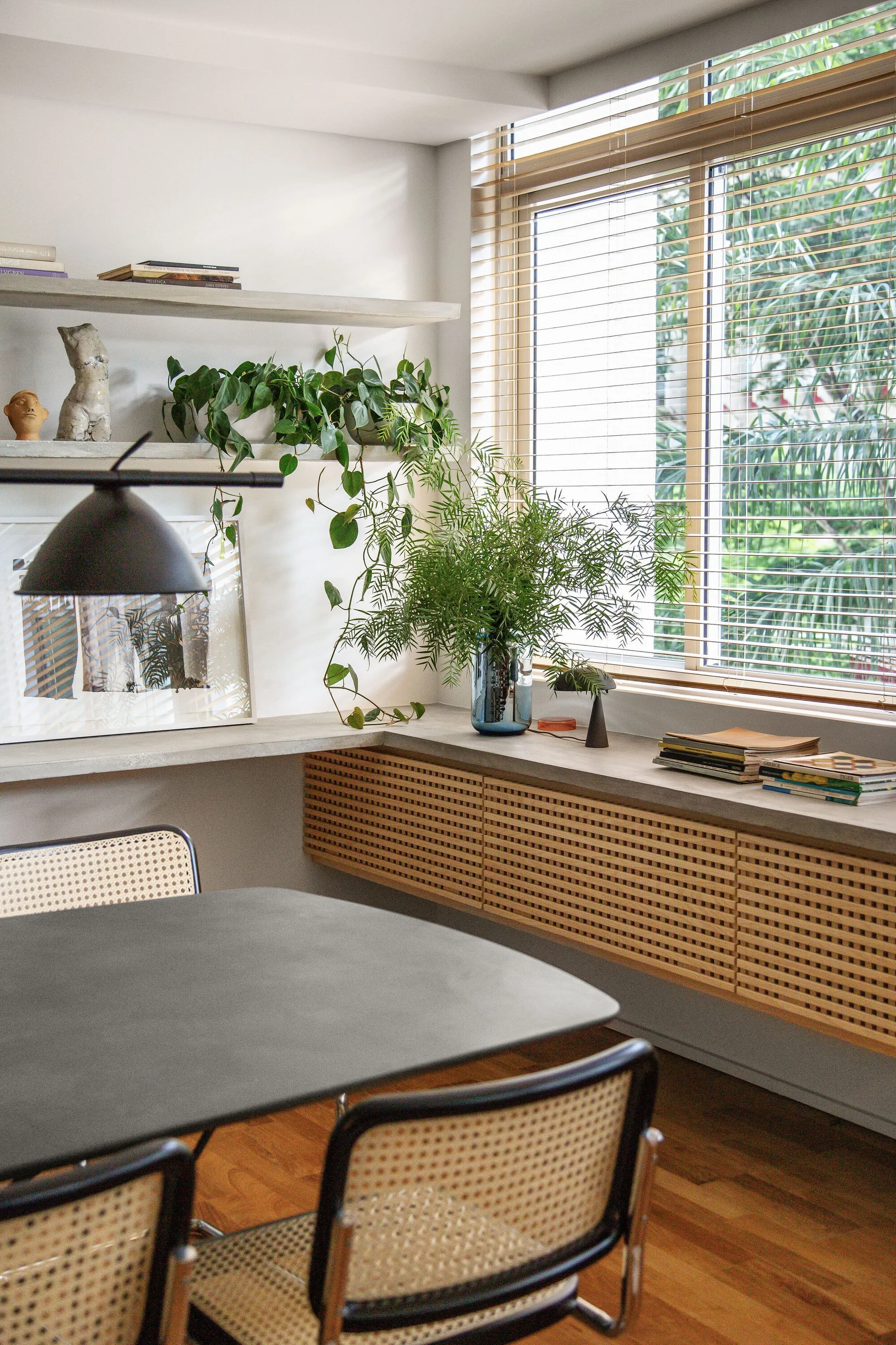 A dining room features cane-front cabinets, concrete-look floating shelves with plants, and cane chairs around a dark table.