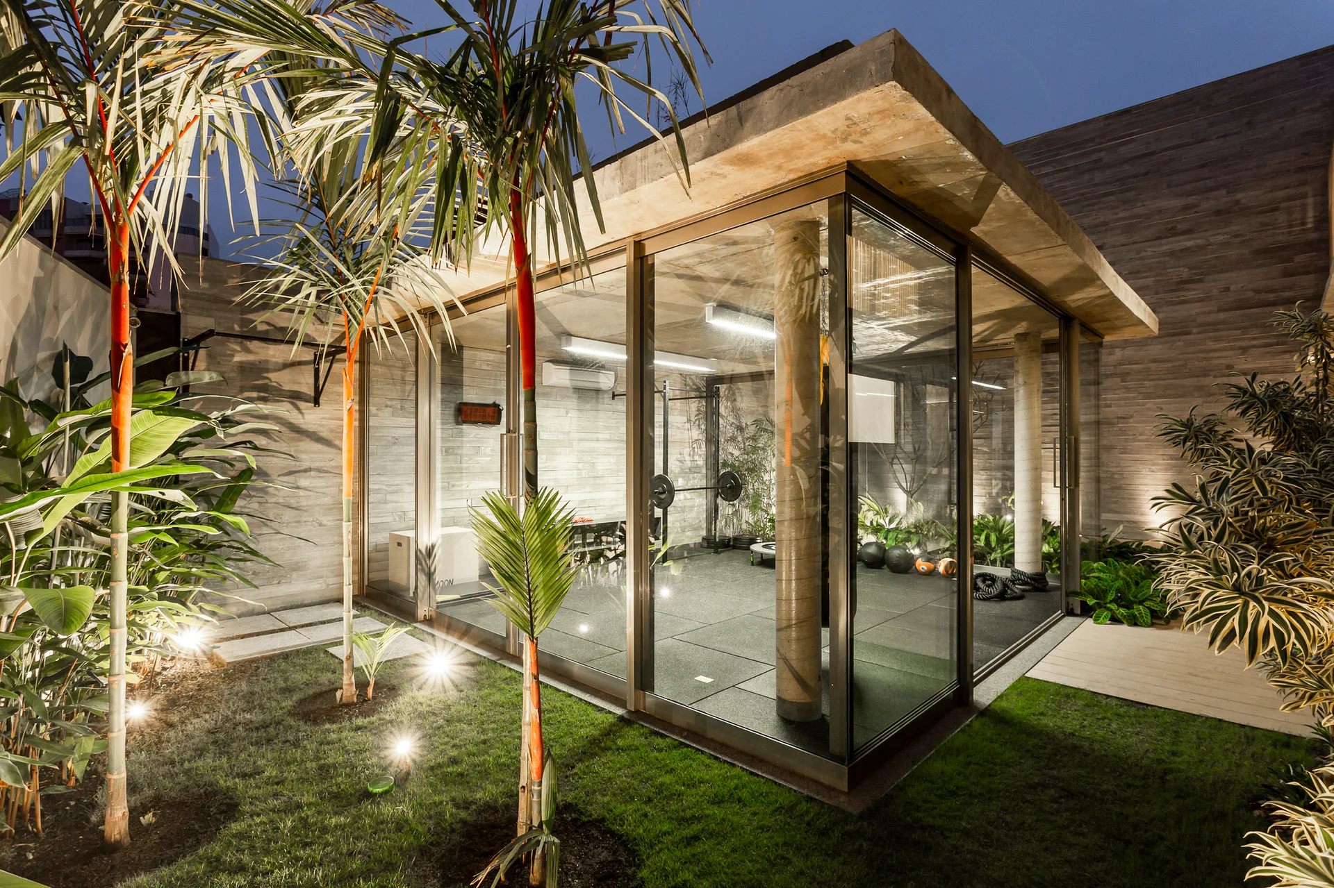 A modern glass-enclosed home gym pavilion with a concrete roof sits in a landscaped garden with palm trees and ambient lighting.