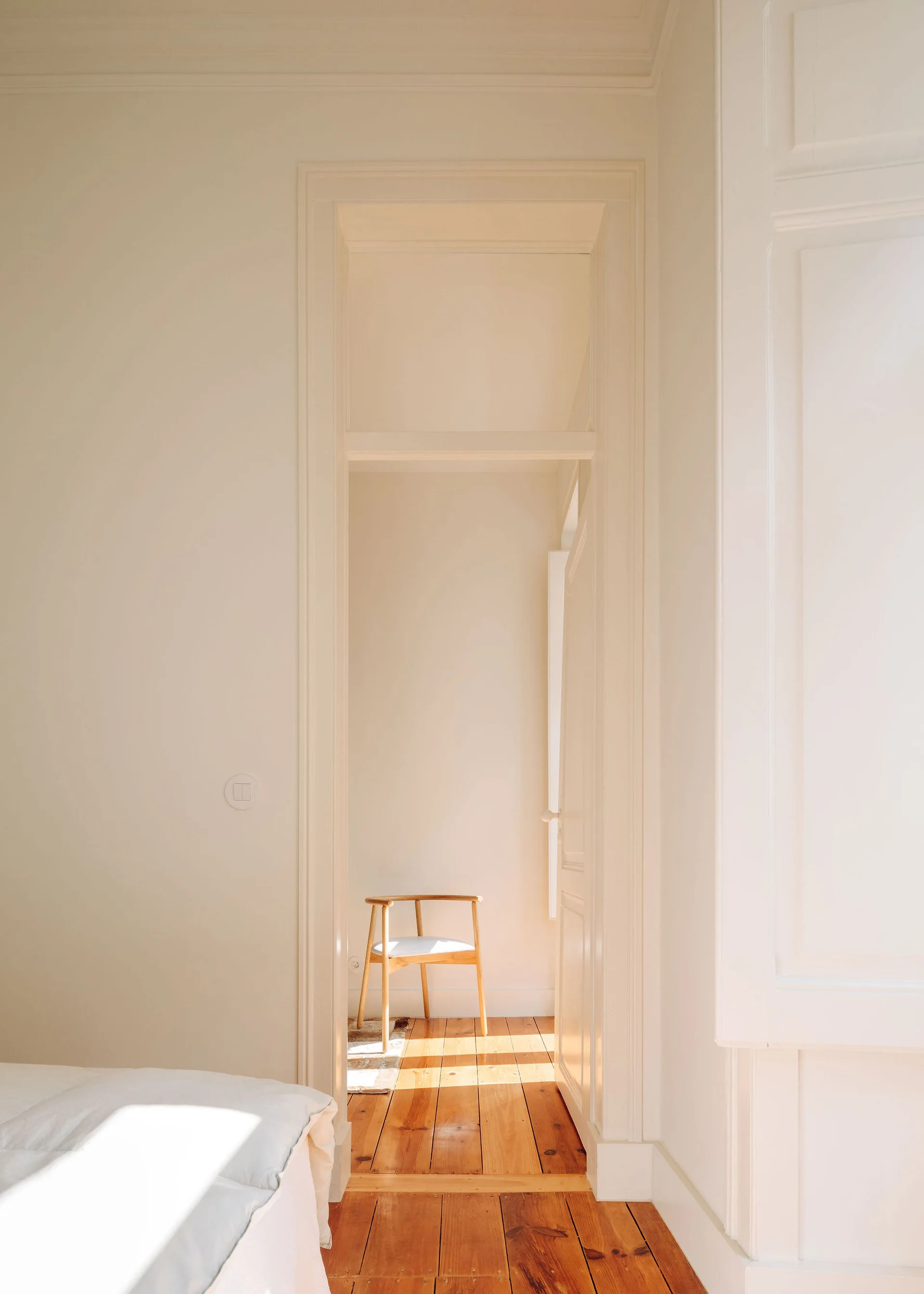 A view through an open doorway from a bedroom with white bedding to a sunlit adjoining room featuring warm wooden floors and a single wooden chair.