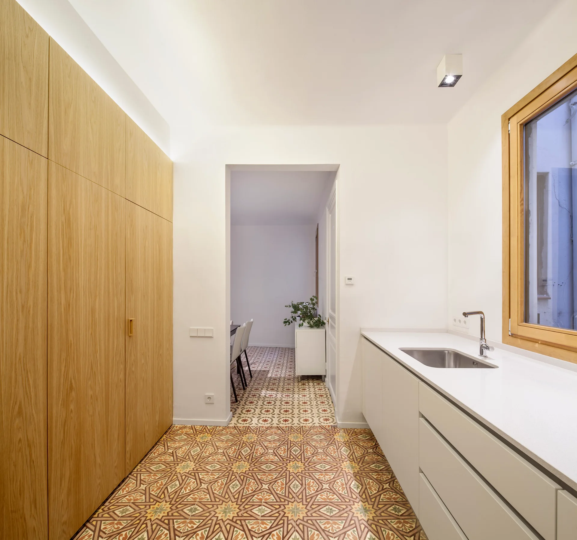 A minimalist kitchen features tall wood cabinetry, white base cabinets with an integrated sink, and intricate patterned floor tiles.