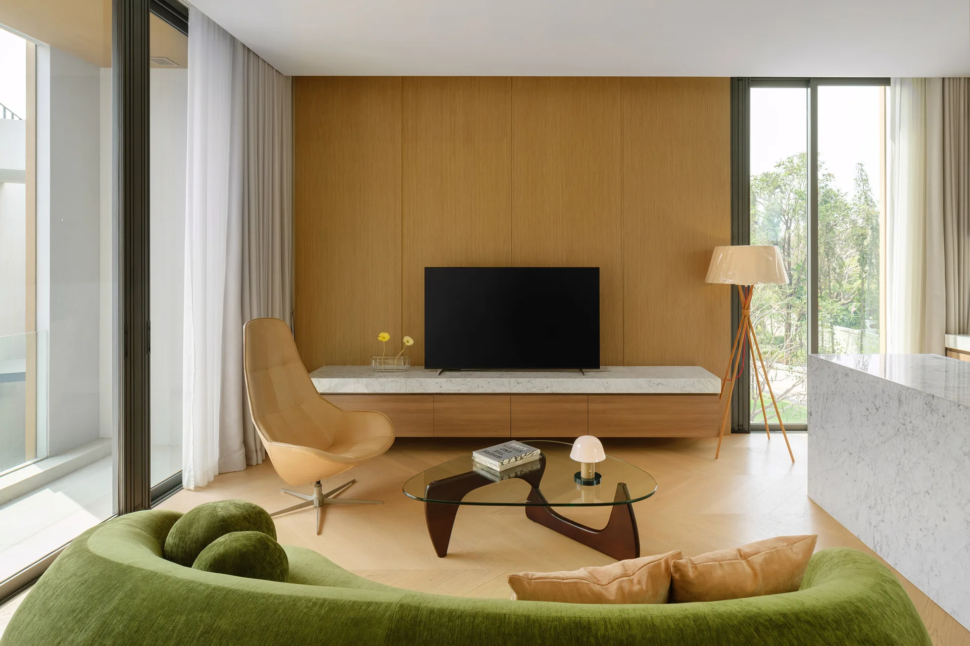 A modern living room featuring a green curved sofa, glass coffee table, and a tan leather armchair in front of a wood-paneled wall with a TV console.