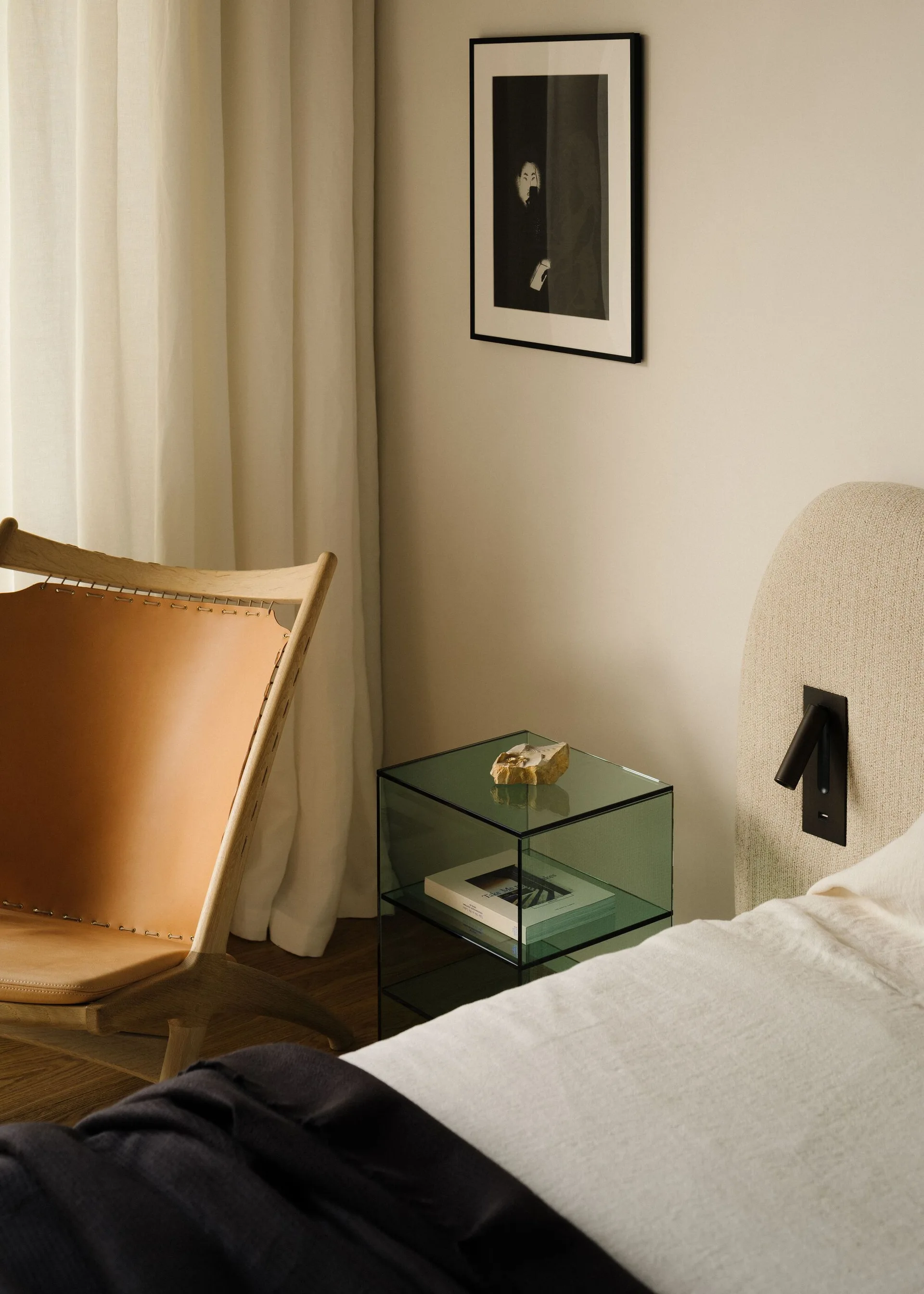 A minimalist bedroom featuring a textured beige headboard, a green tinted glass nightstand, and a tan leather accent chair.