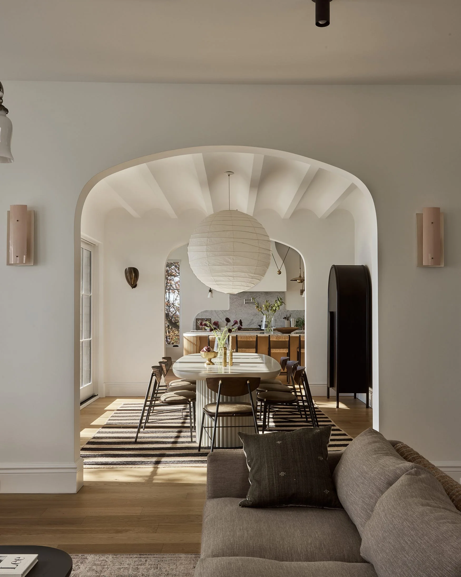 A view through a plaster archway into a dining room featuring an oval table, striped rug, and large paper pendant light.