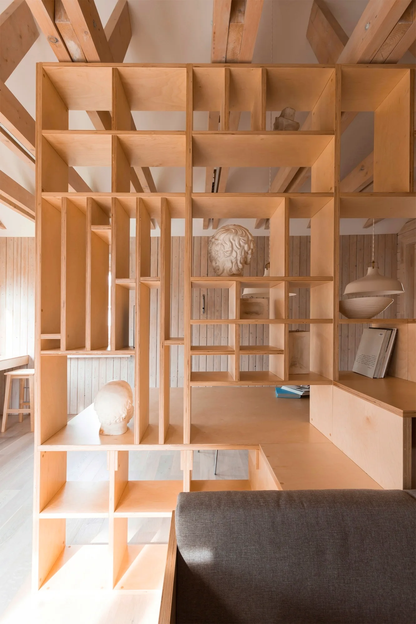 A custom plywood open shelving unit serves as a room divider next to a gray sofa under exposed timber ceiling beams.