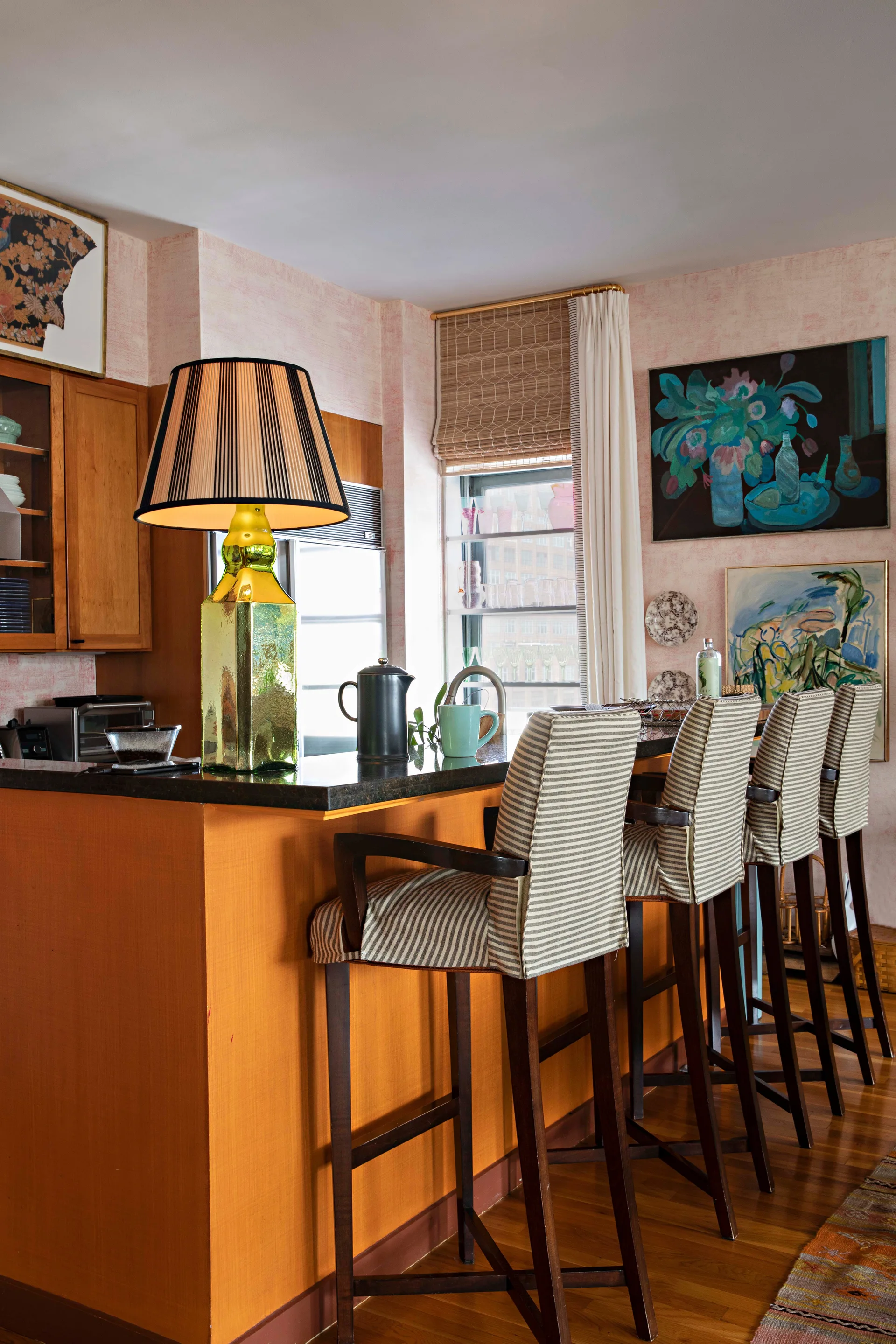 A vibrant kitchen space featuring a bright orange island with a black counter, striped high-back bar stools, and textured pink wallpaper.