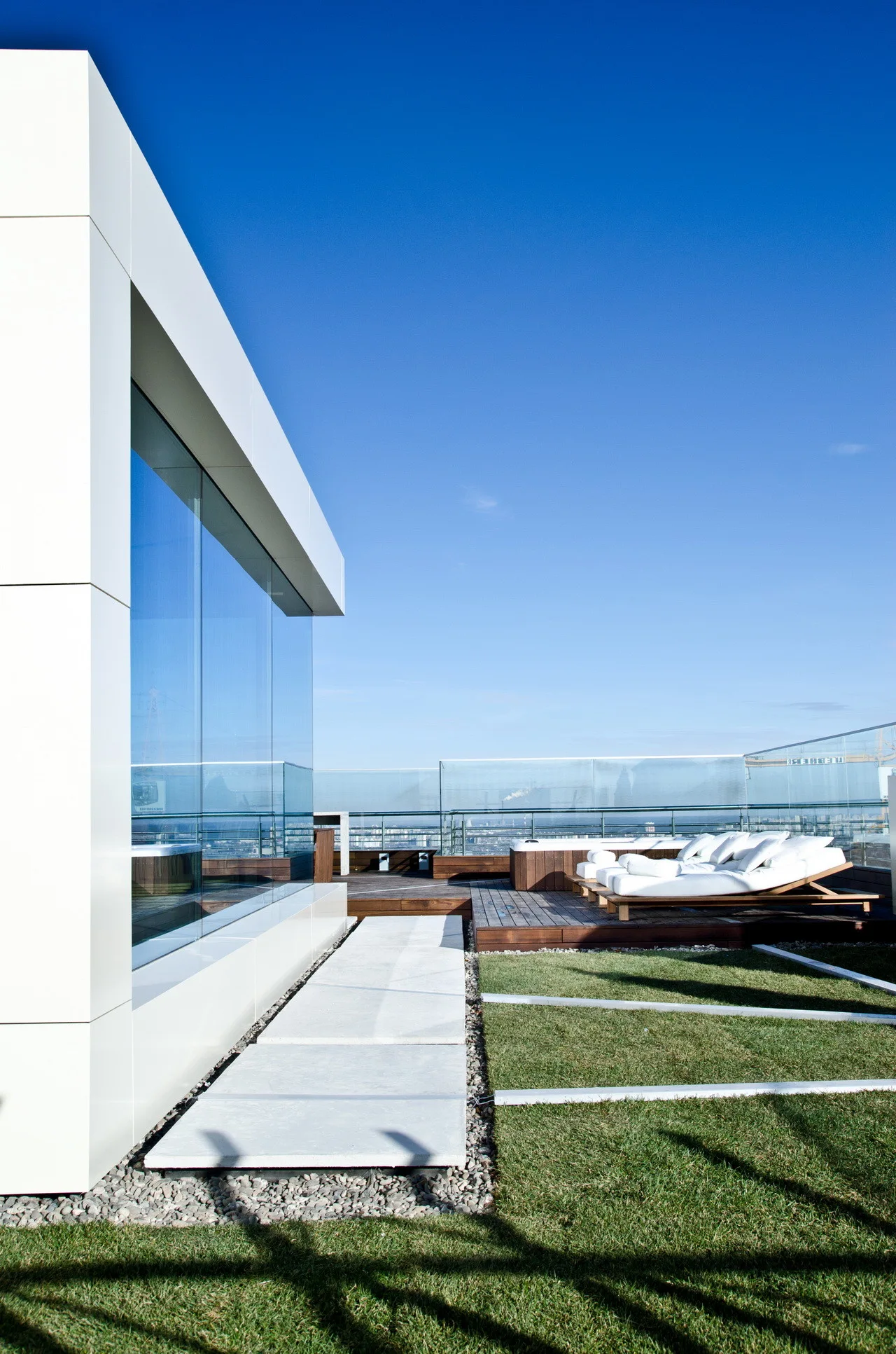 A modern rooftop terrace featuring a wooden deck with white lounge chairs, an integrated grass lawn, concrete pavers, and a glass perimeter railing.