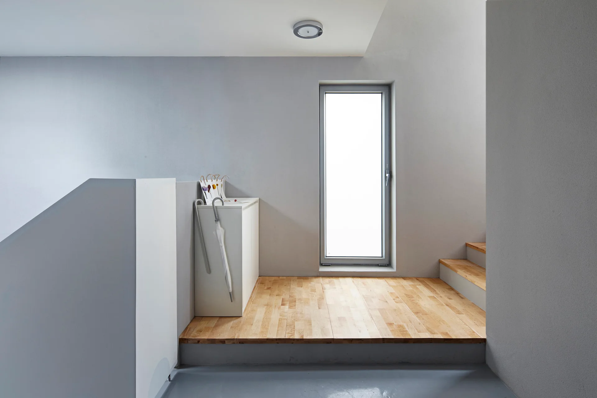 A minimalist stair landing features a raised light wood platform, grey walls, a tall narrow window, and wooden stairs on the right.
