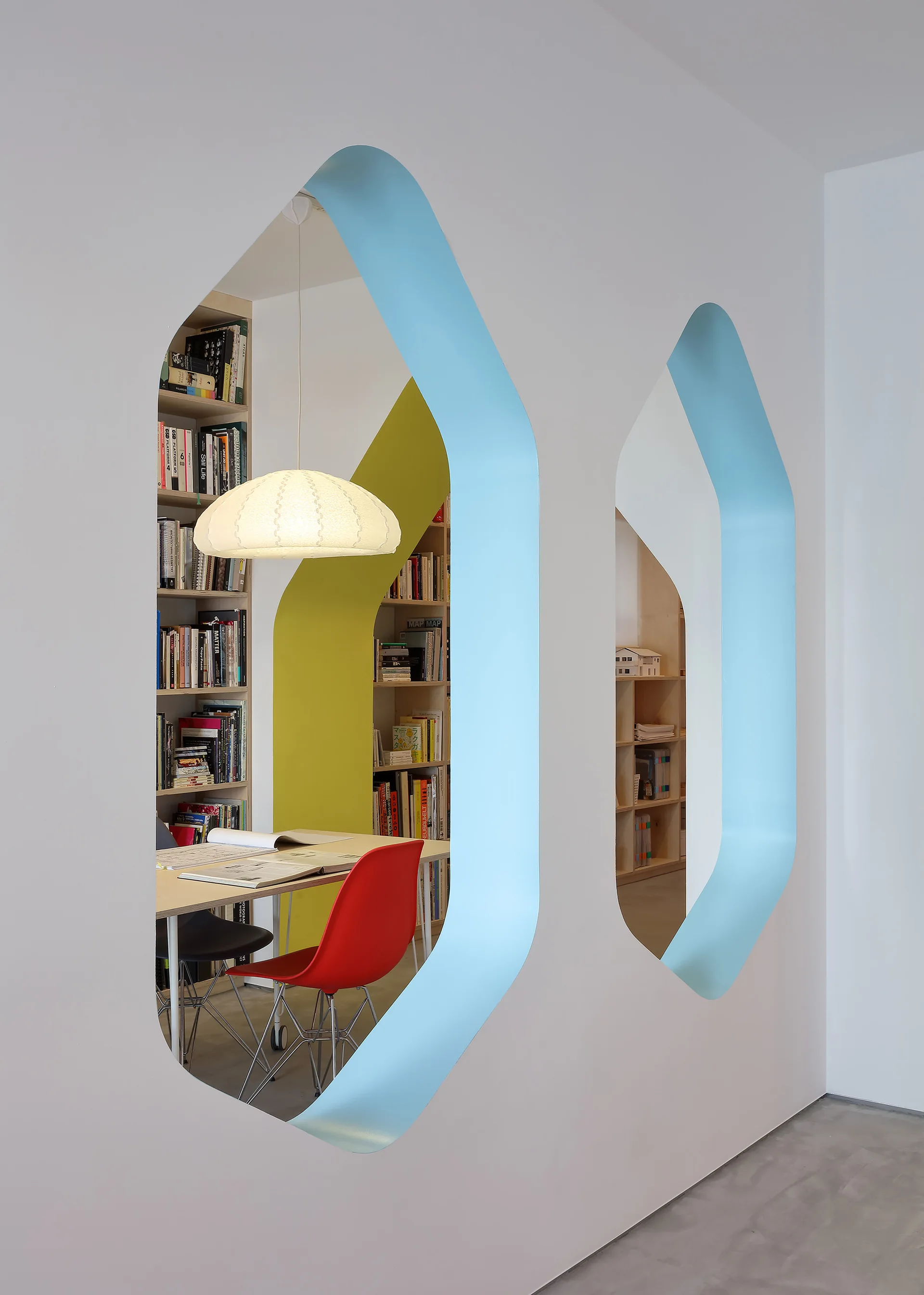 A white wall with light blue geometric cutouts reveals a workspace with wooden bookshelves, a desk, a textured pendant lamp, and a red chair.