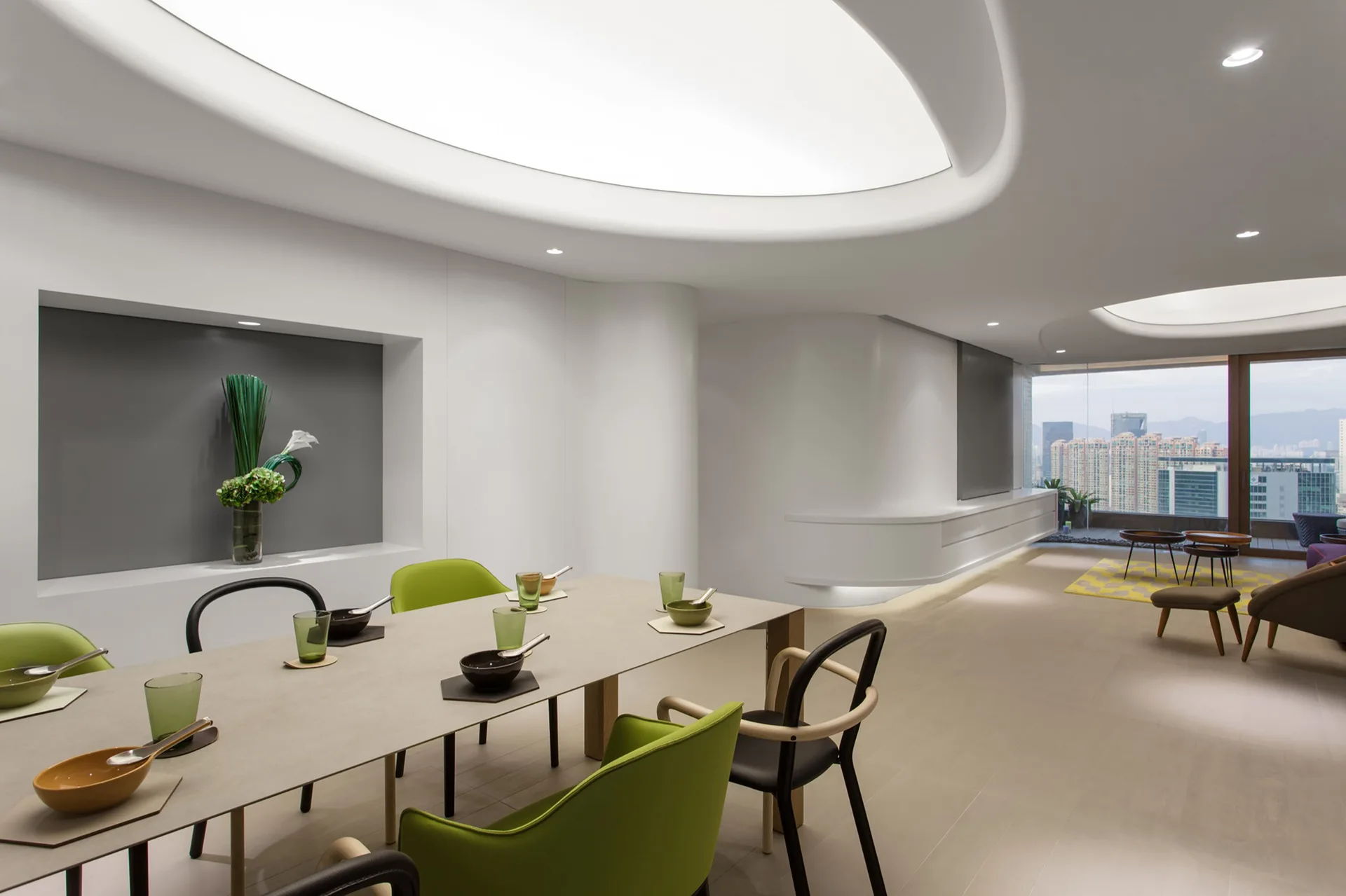 A modern open-plan dining room features a long table, vibrant green chairs, a gray recessed wall niche, and large oval illuminated ceiling coves.