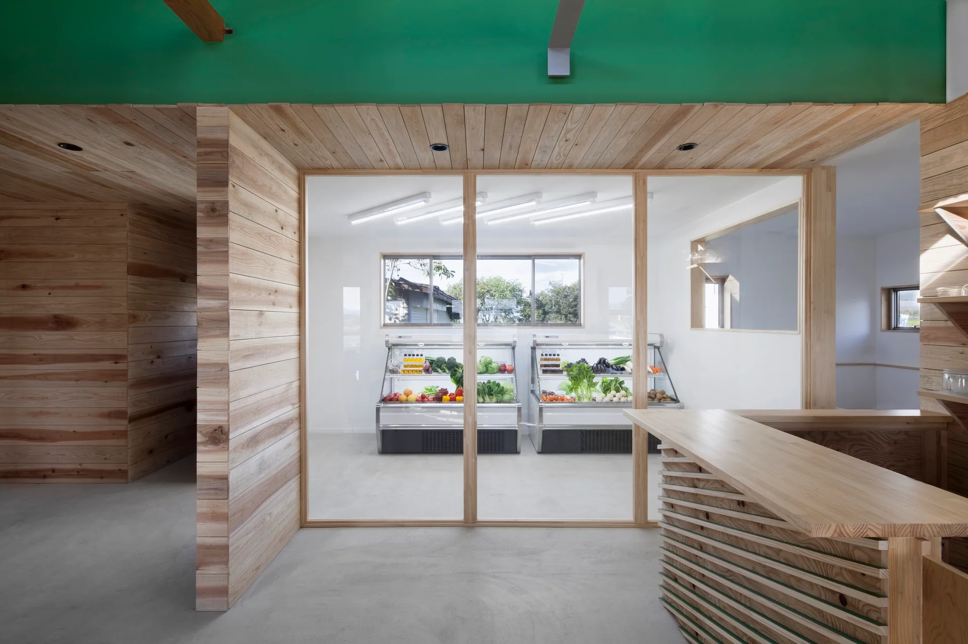A minimalist retail interior with light wood paneled walls, a large glass partition, and refrigerated produce displays in the back.