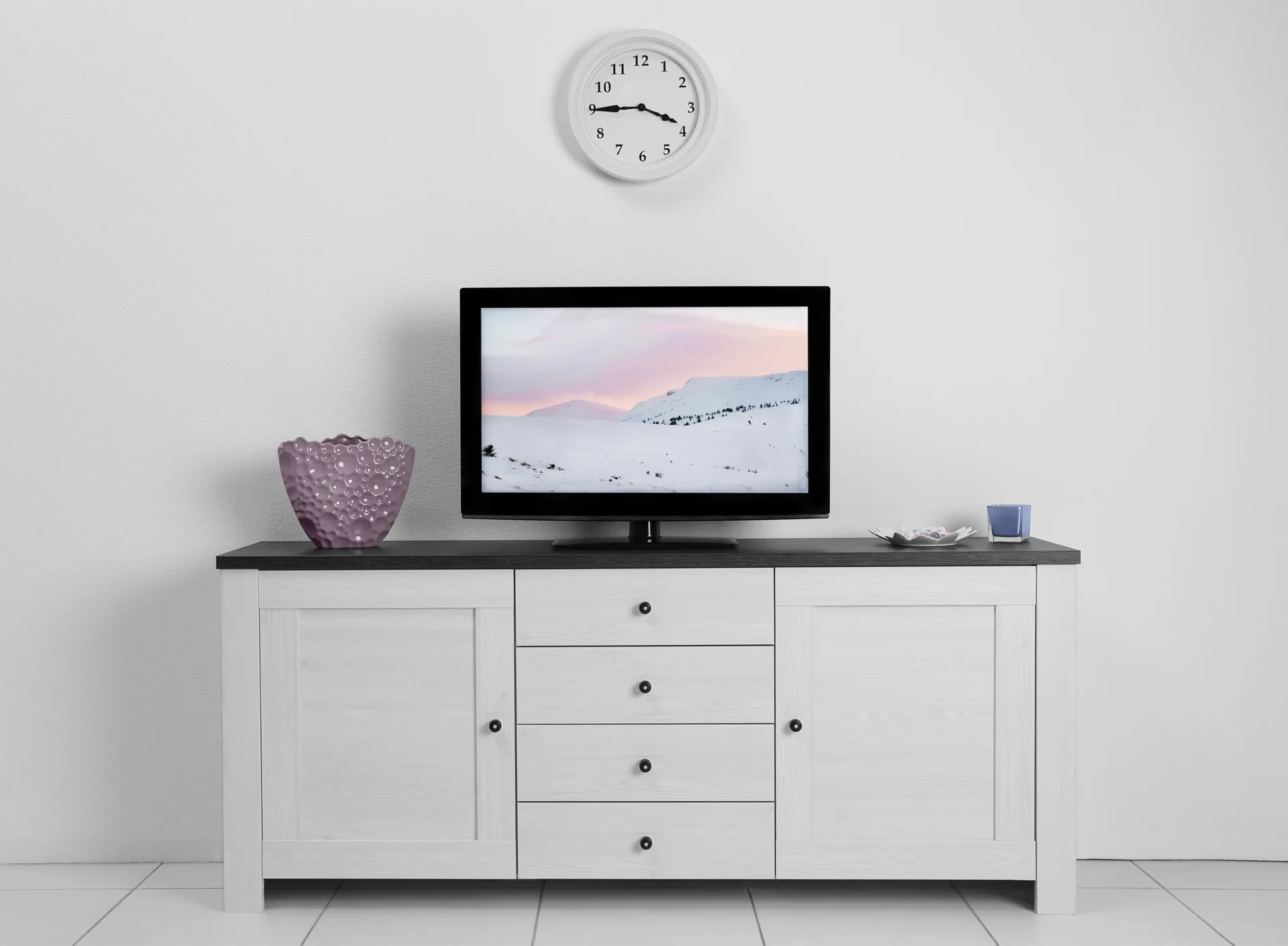 A white media console with a black top holds a television, a textured purple vase, and a small blue candle holder against a white wall.
