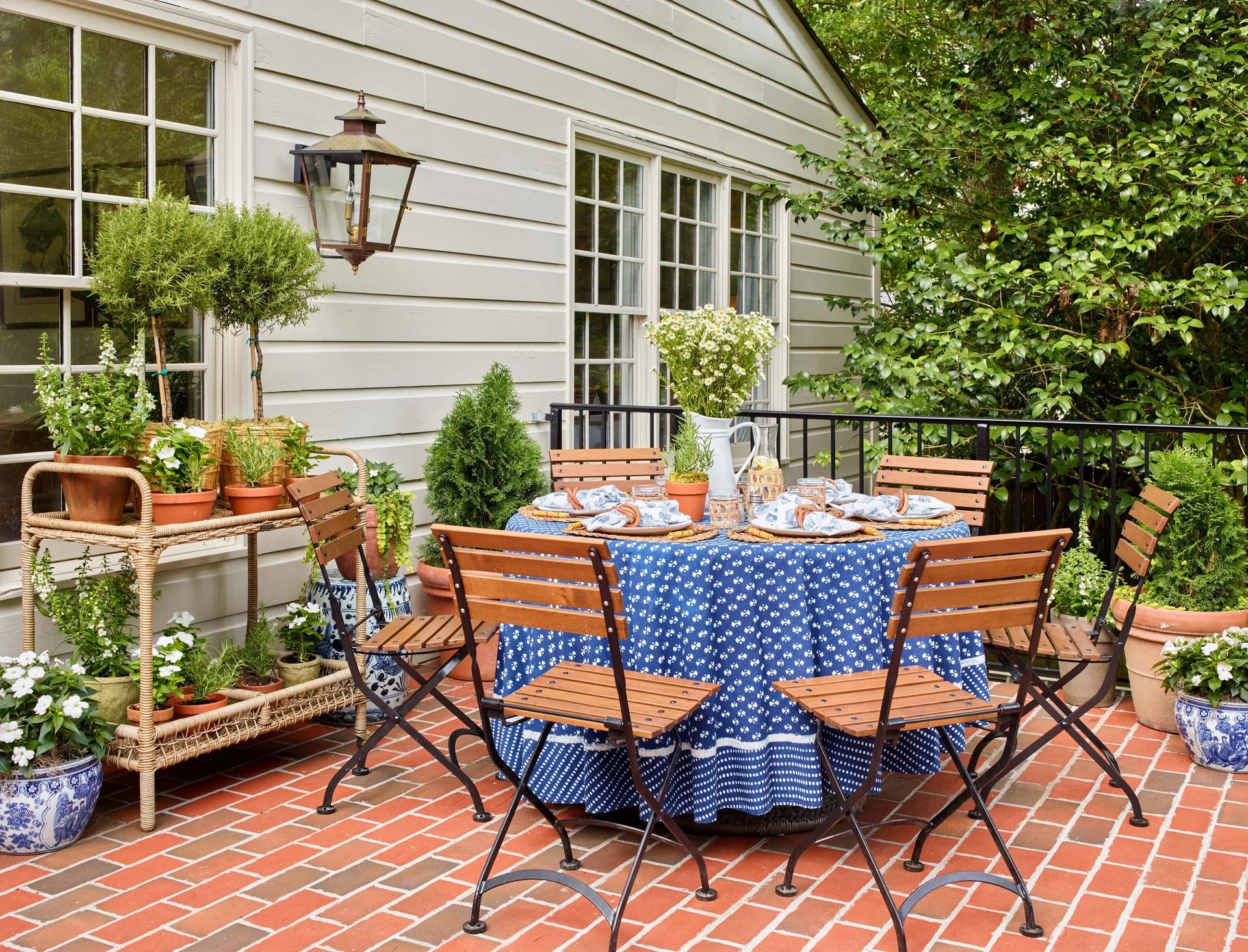 An outdoor dining table with a blue patterned tablecloth and wood-slatted chairs sits on a red brick patio next to a house with gray siding.
