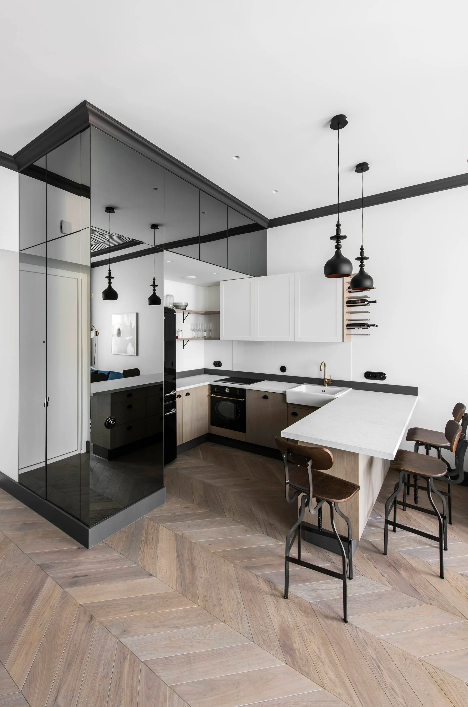 A modern kitchen features light wood cabinets, a mirrored wall panel, herringbone wood floors, and wooden bar stools at a white peninsula.