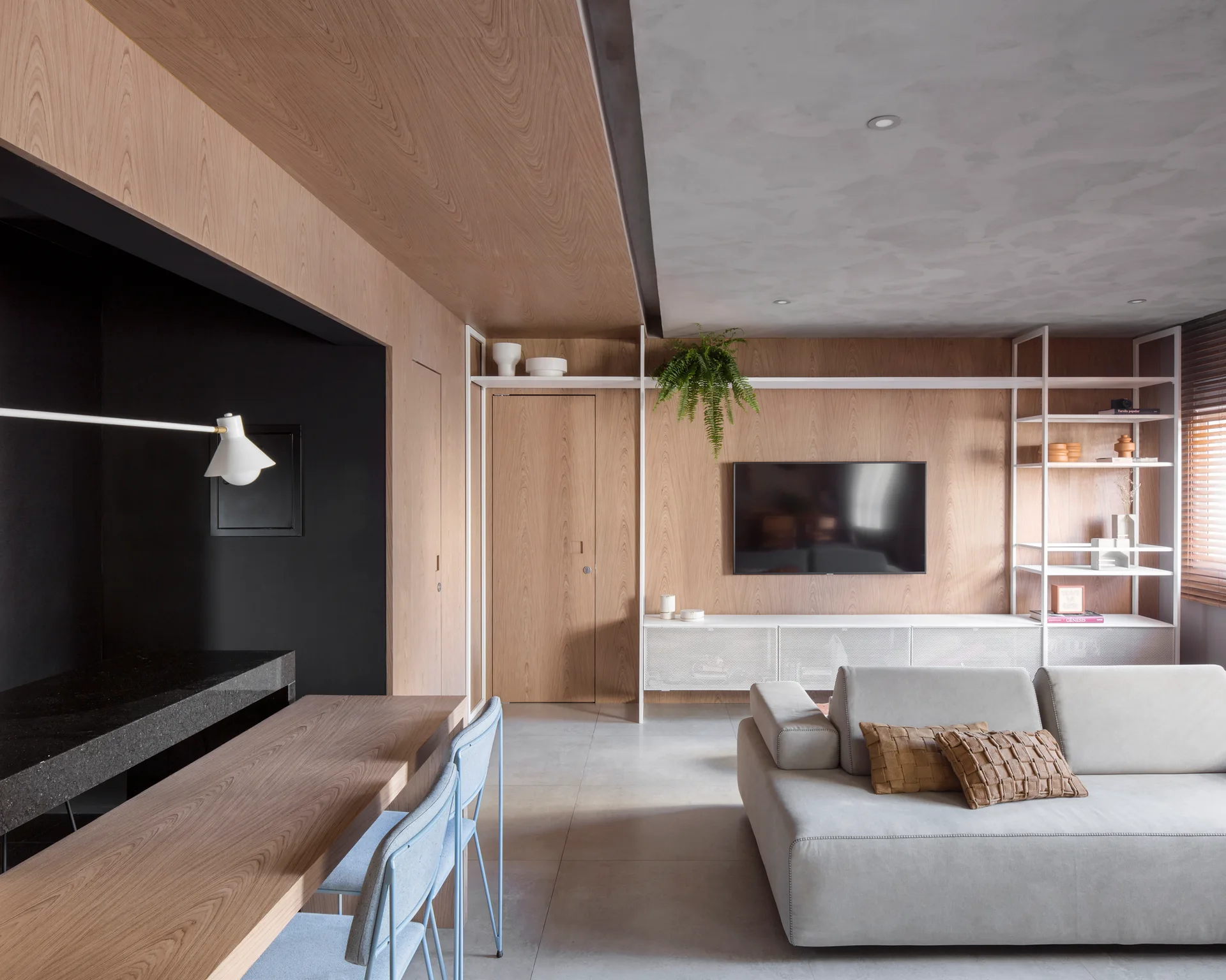 A contemporary living and dining area features a light gray sofa, custom wood veneer wall paneling, and white metal open shelving framing a television.