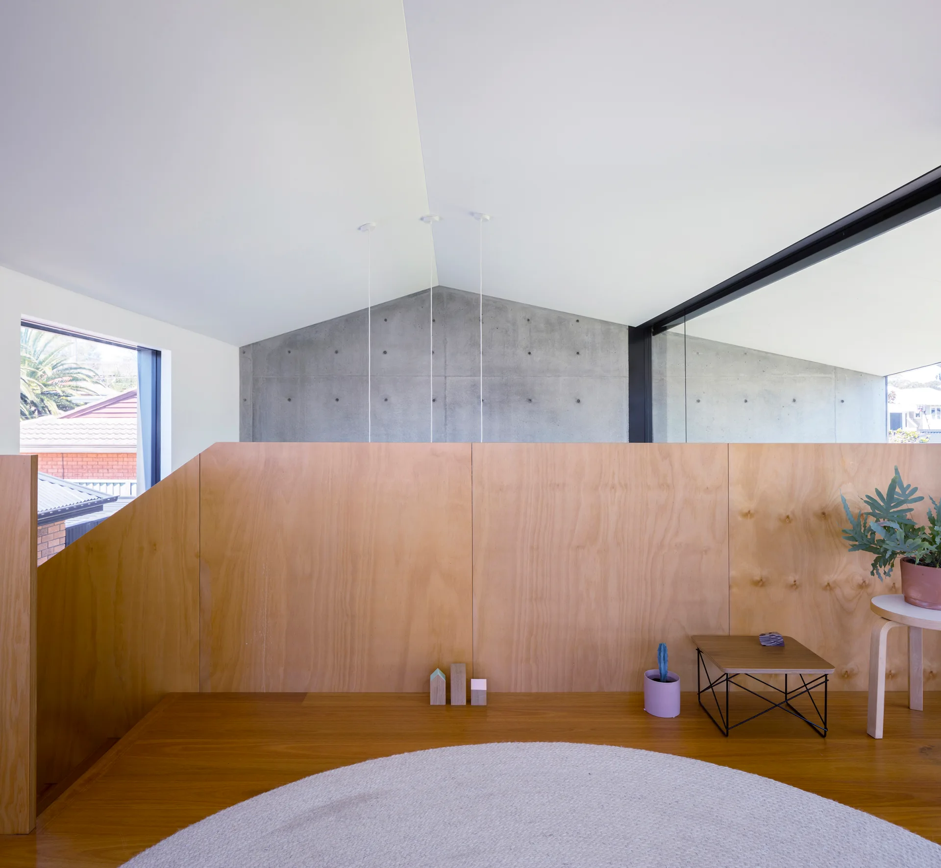 A minimalist loft landing features a plywood partition wall, wooden flooring, an angled white ceiling, and a grey concrete wall in the background.