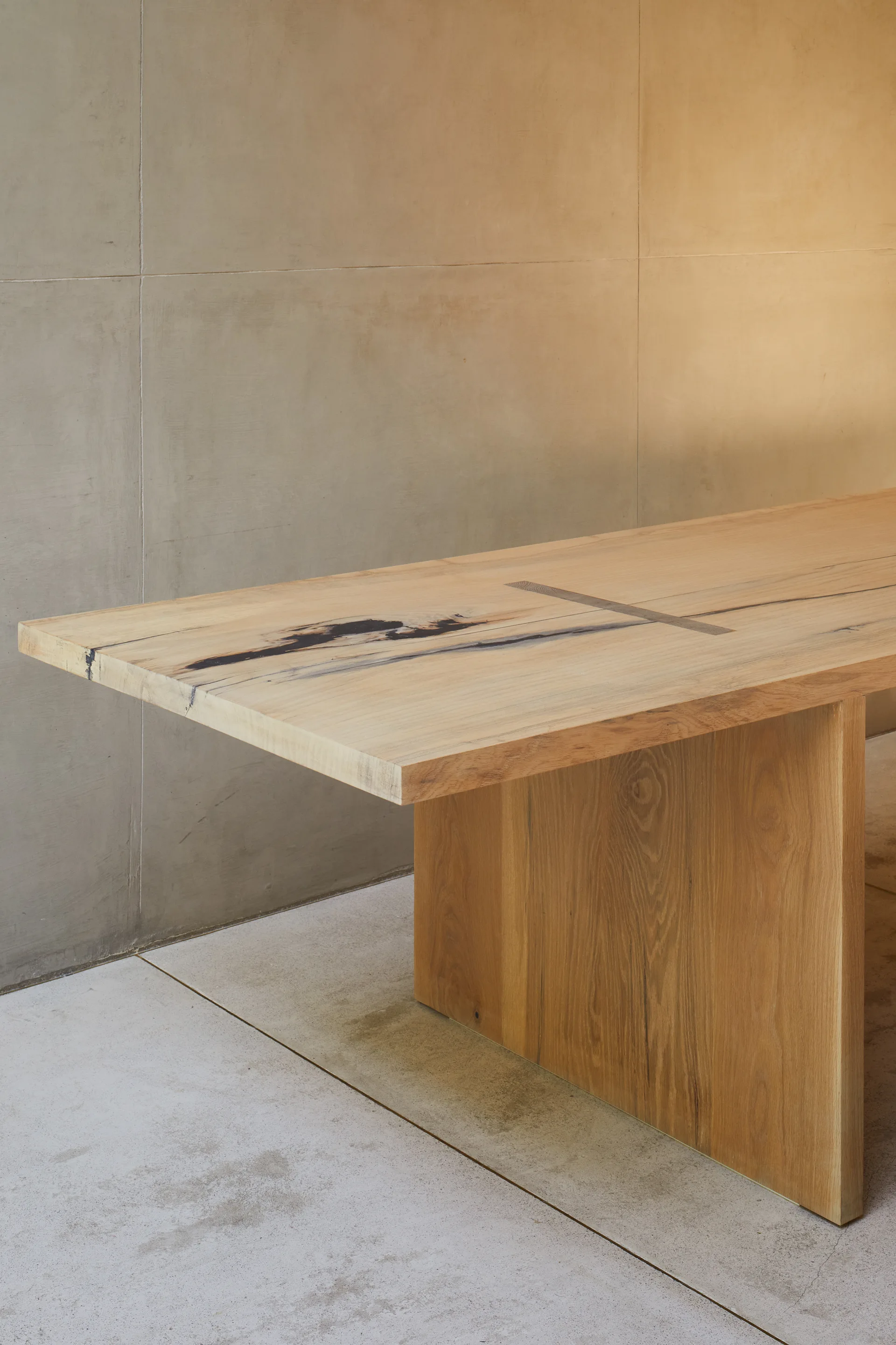 A light-toned solid wood dining table with a slab leg is positioned against a smooth concrete panel wall and concrete floor.