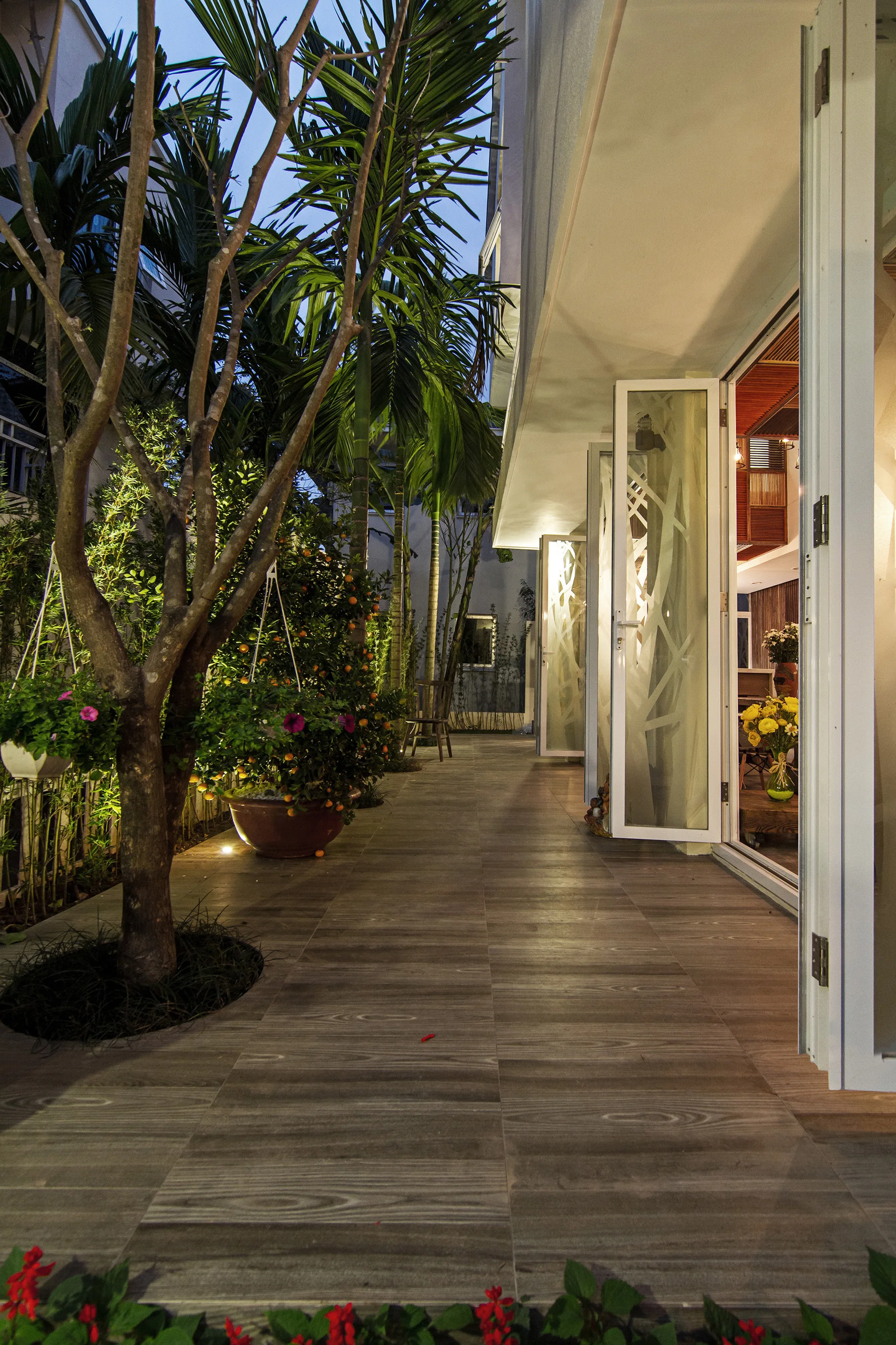 An outdoor tiled walkway at dusk with a tree growing from a floor cutout next to open folding glass doors revealing a lit interior.