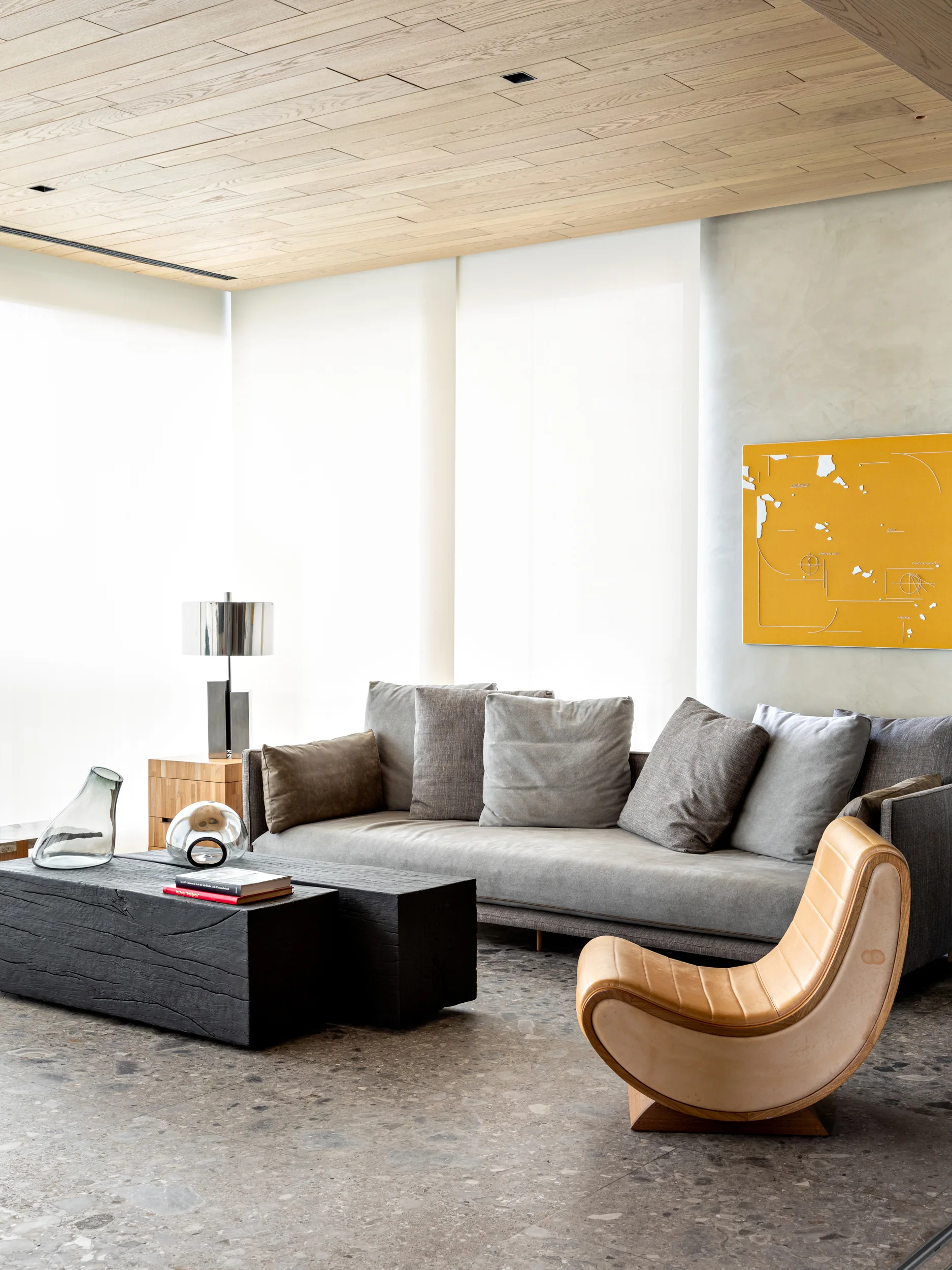 A contemporary living room features a grey sofa, black block coffee table, and a curved tan leather lounge chair beneath a light wood plank ceiling.