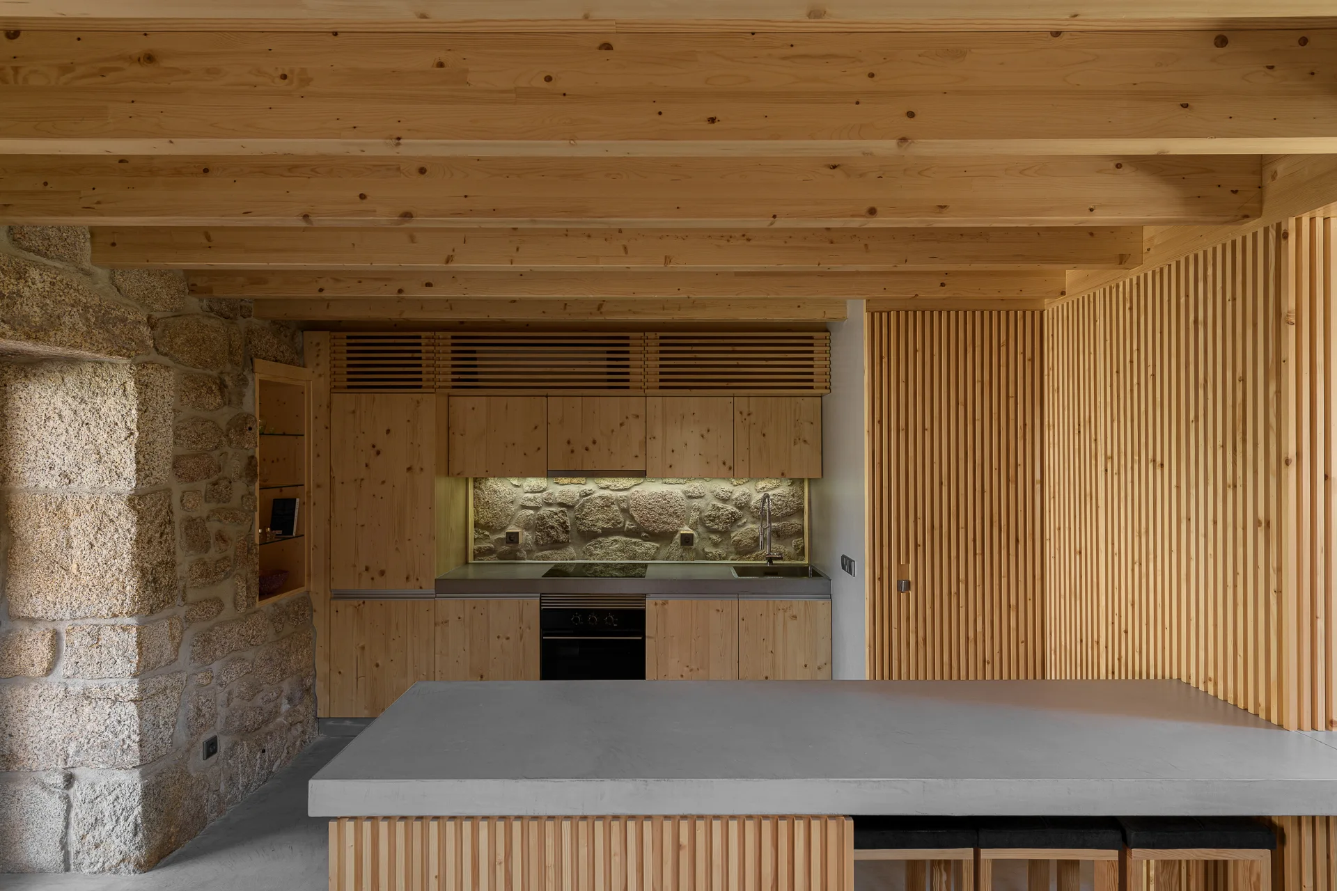 A modern rustic kitchen featuring flat-front light pine cabinetry, an exposed stone wall, a gray concrete island, and a timber-beamed ceiling.