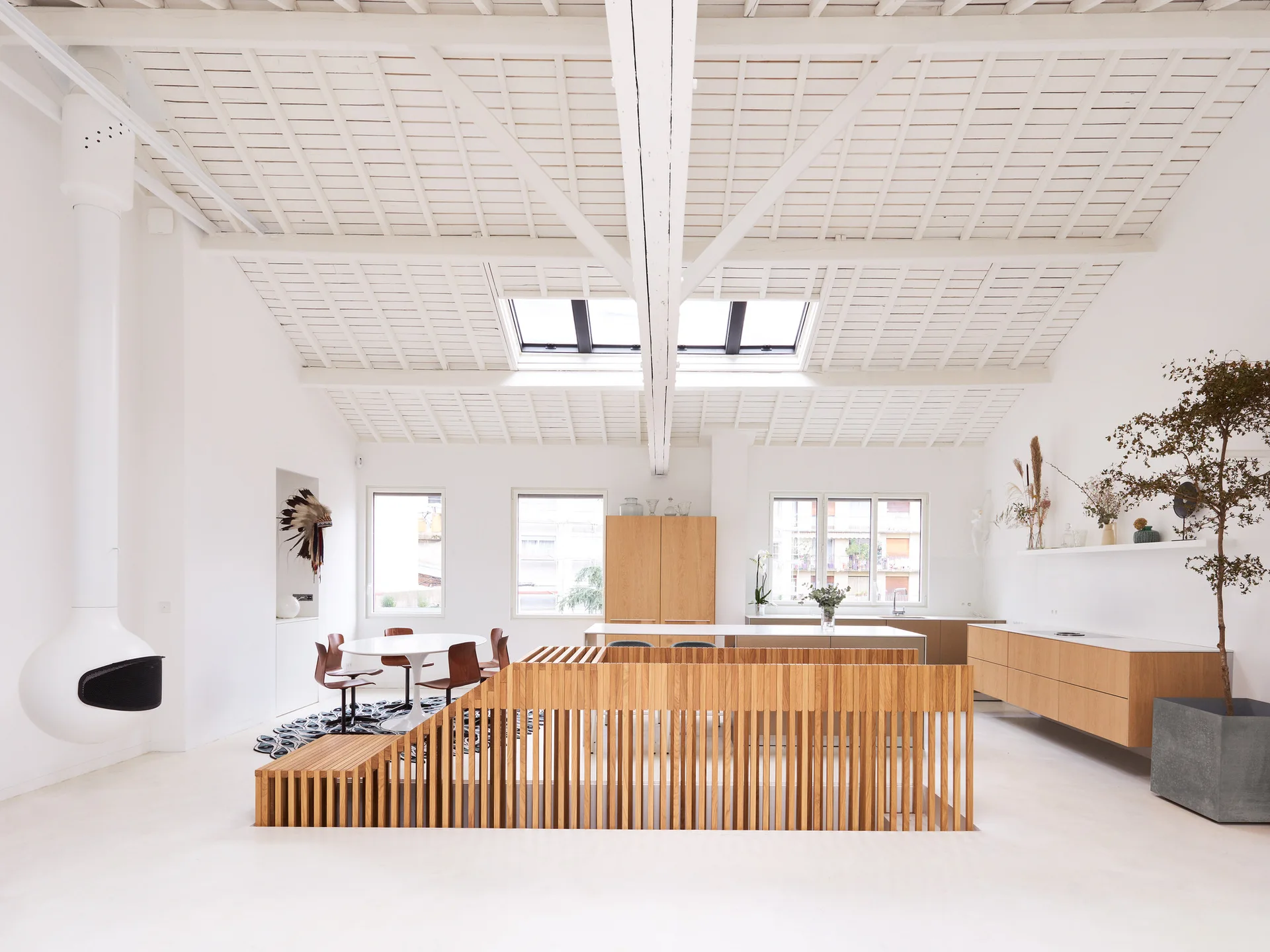 An open loft space with a stepped slatted wood railing, white hanging fireplace, and wood cabinetry under a beamed ceiling with skylights.