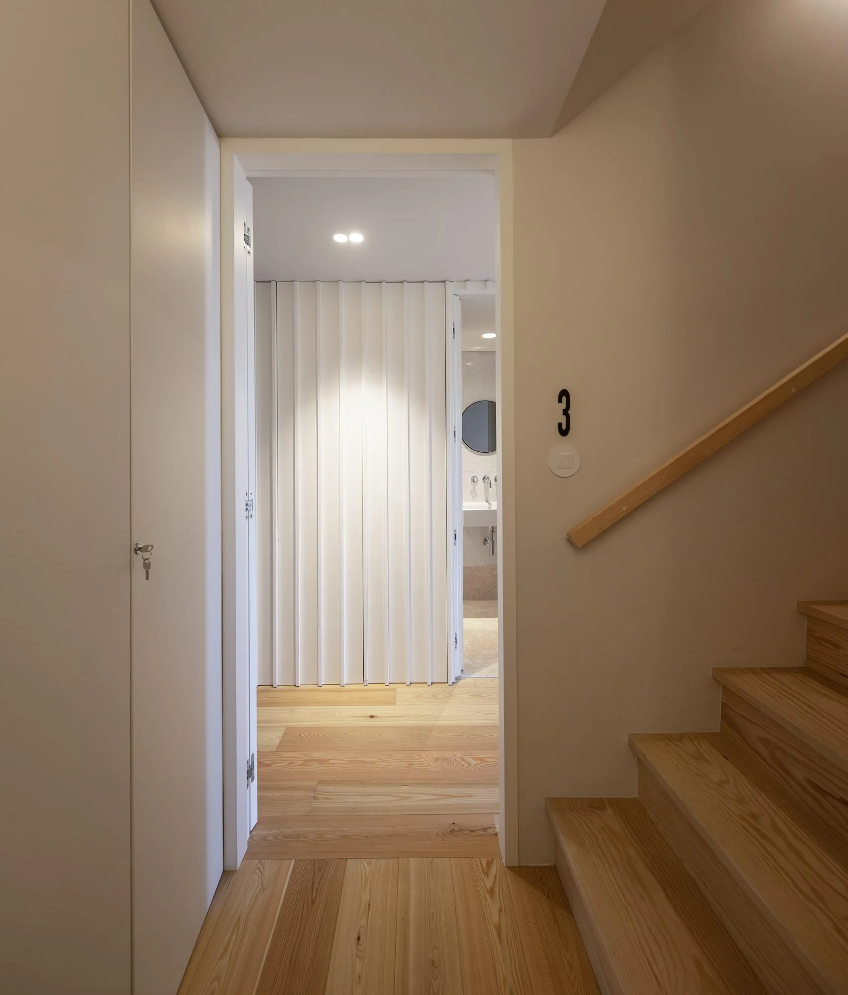 A minimalist hallway showcases light wood stairs on the right, a matching wooden handrail, and an open doorway leading to a white partitioned bathroom.