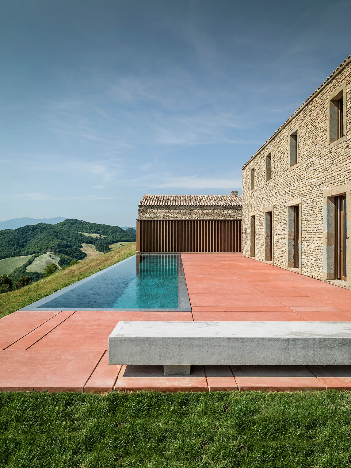 A modern infinity pool on a terracotta terrace next to a stacked stone building, featuring a concrete bench and a scenic hill view.