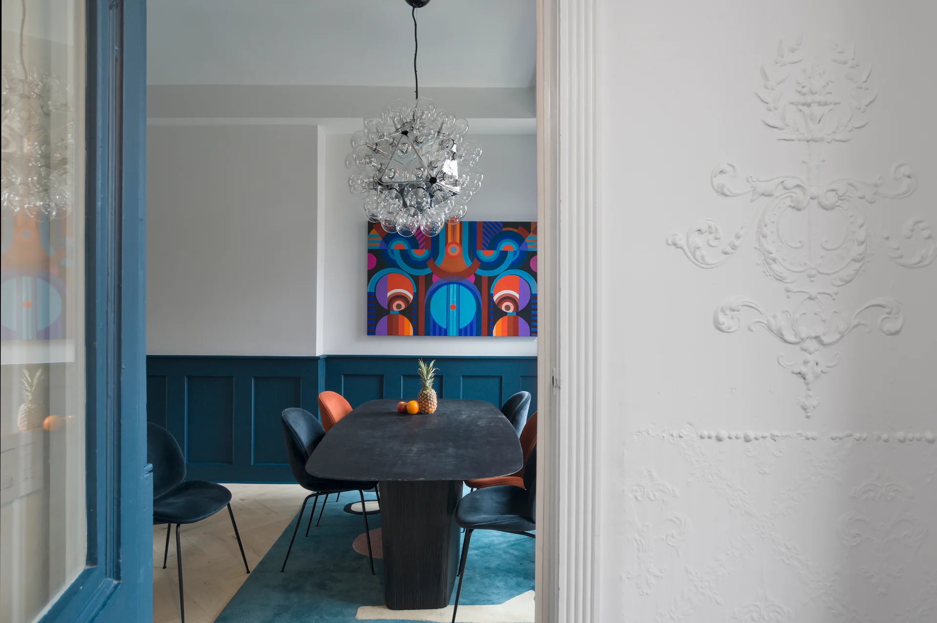 A modern dining room seen through a white doorway features a black table, blue velvet chairs, deep blue wainscoting, and a bubble glass chandelier.