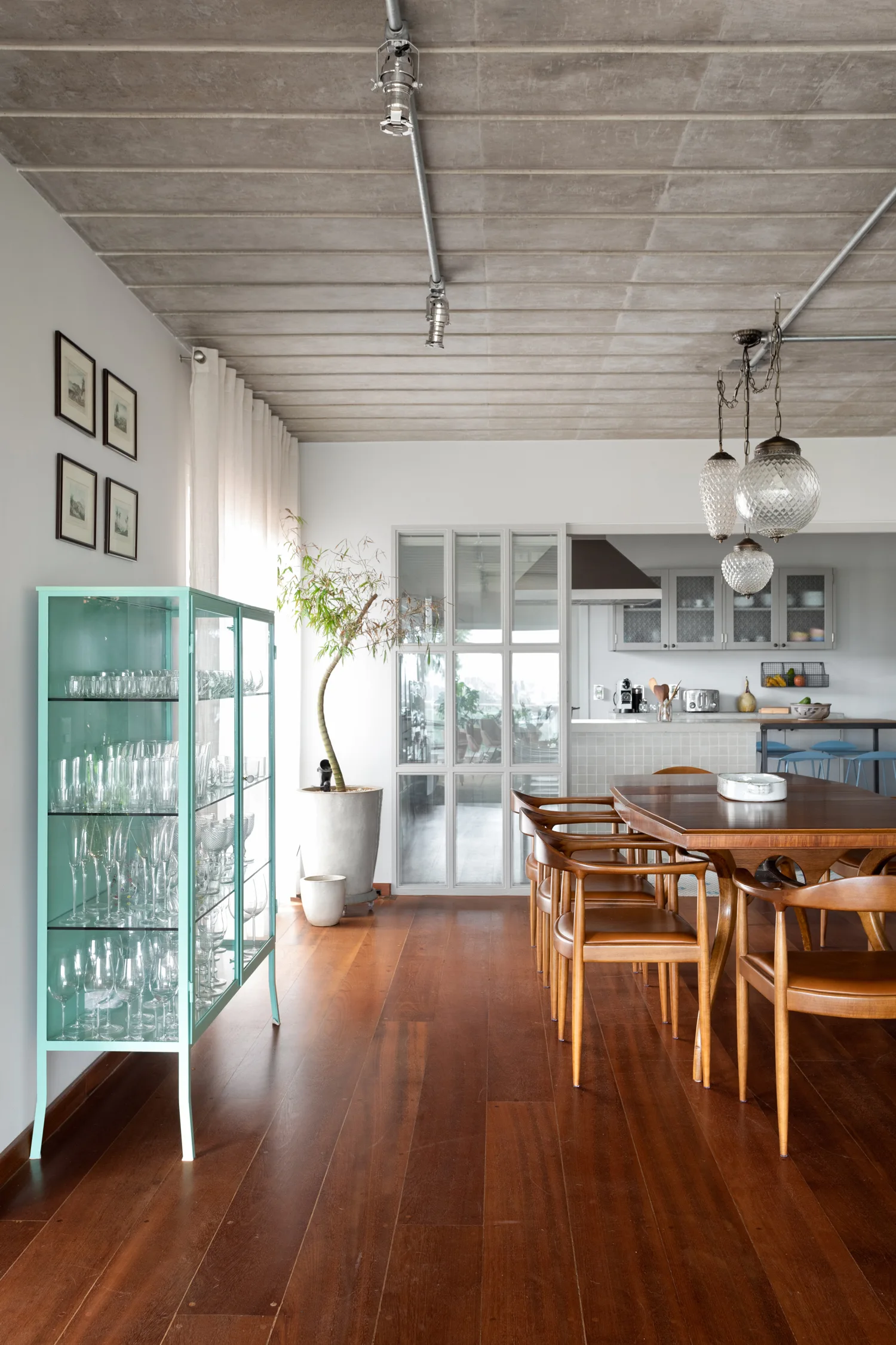 A bright dining room features a wooden table and chairs, an exposed concrete ceiling, and a tall mint green metal display cabinet.