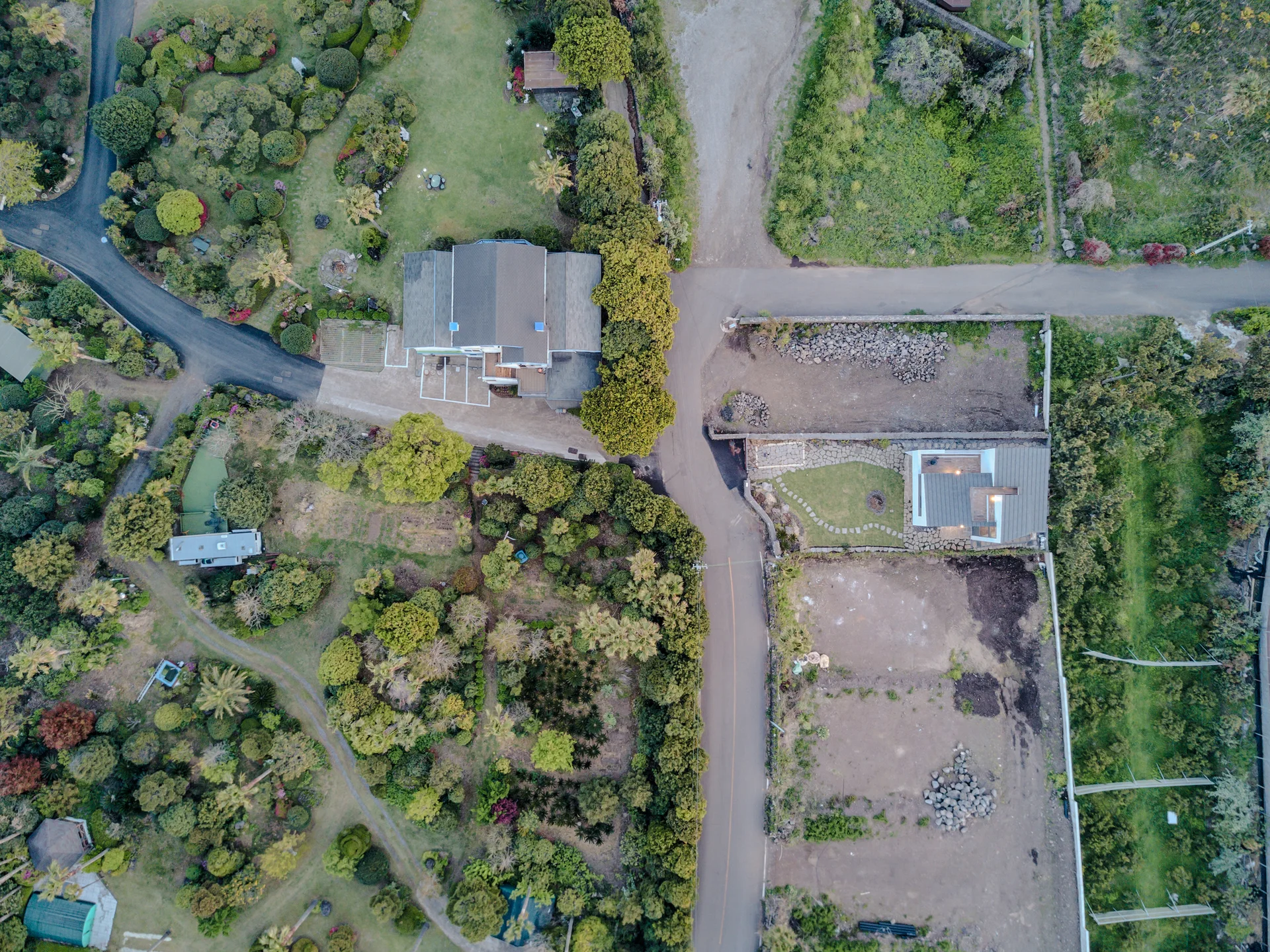 An aerial drone perspective shows residential houses surrounded by lush green gardens, cultivated plots, and stone boundary walls along paved roads.