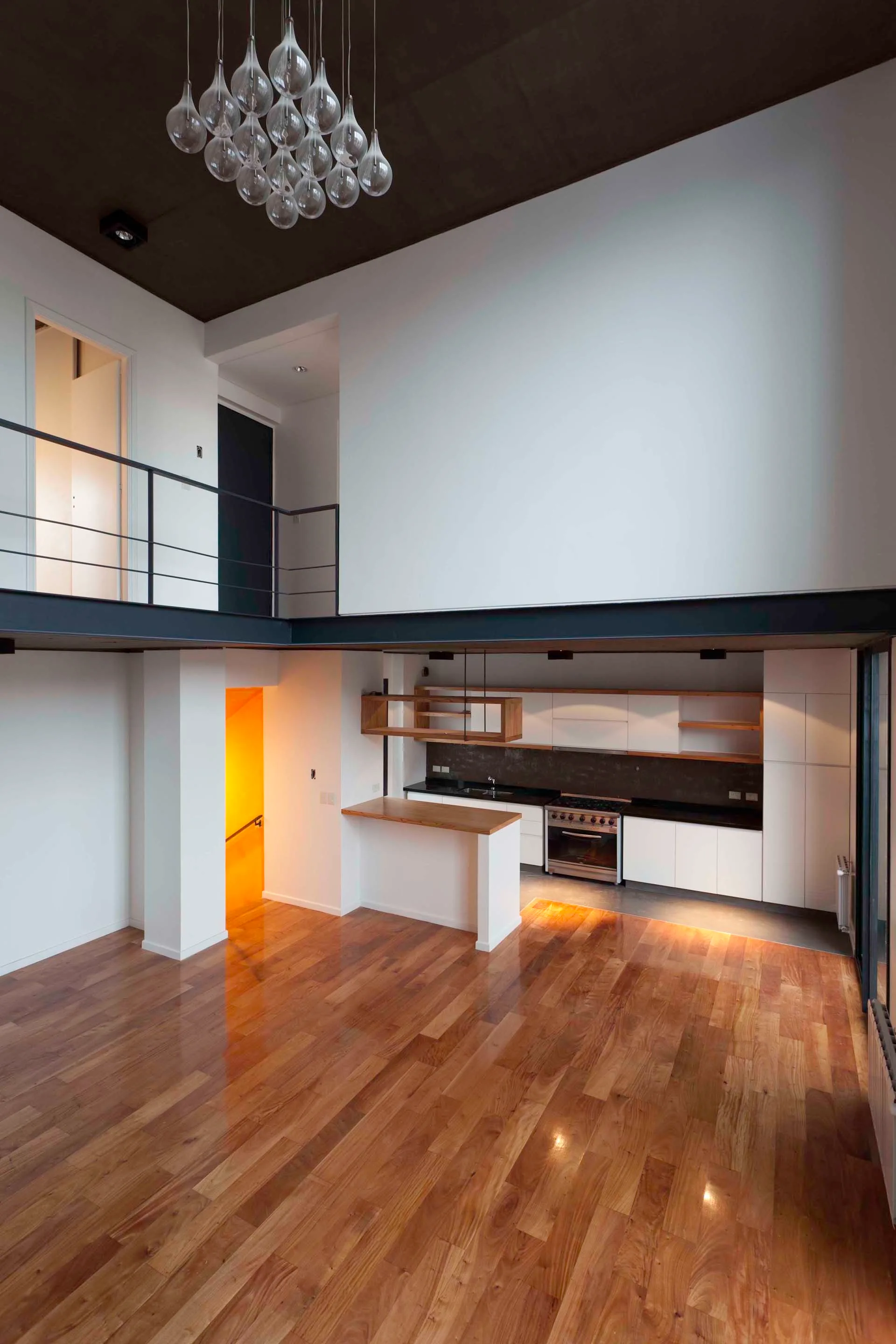 An open-concept living area features polished wood floors, a modern kitchen tucked under a mezzanine level, and a glass cluster chandelier overhead.