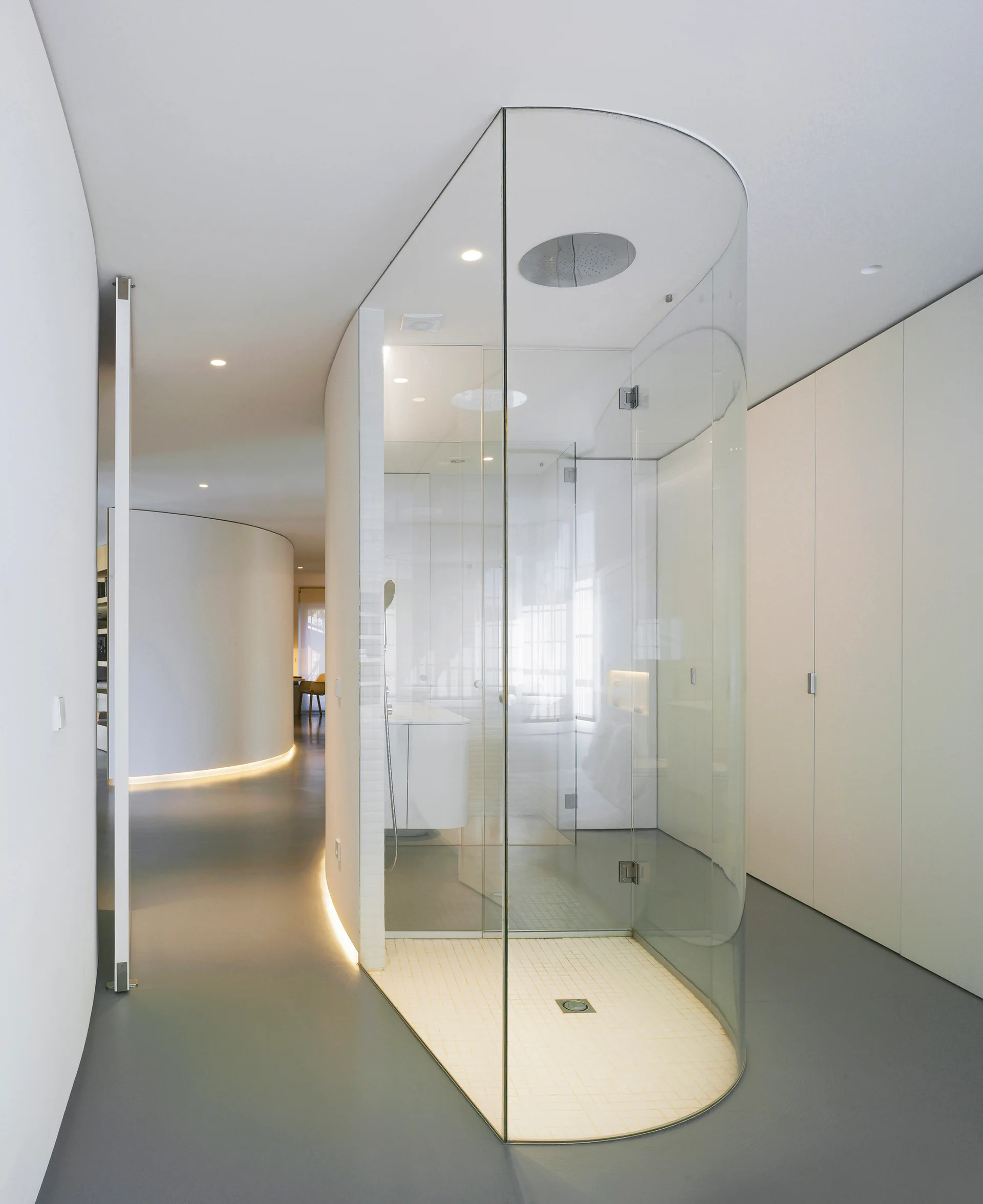 A modern minimalist bathroom featuring a curved glass shower enclosure, smooth white curved walls with floor-level cove lighting, and grey flooring.