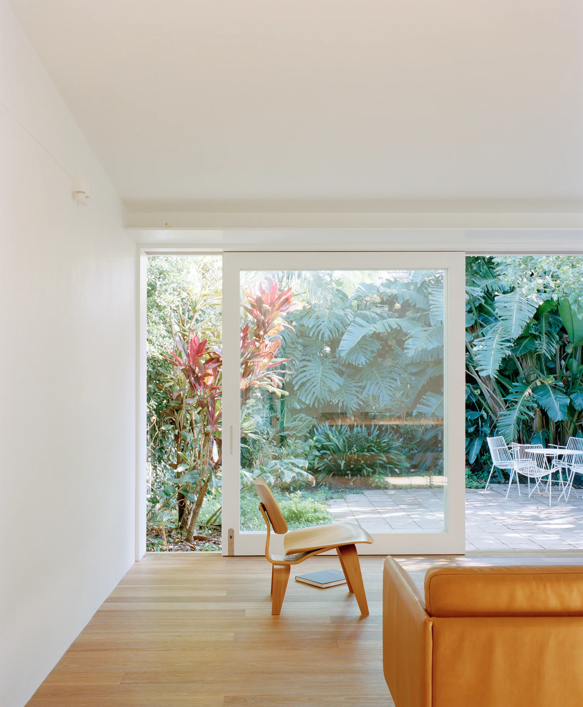A minimalist living room with a tan leather sofa and plywood chair facing sliding glass doors that look out onto a lush green garden patio.