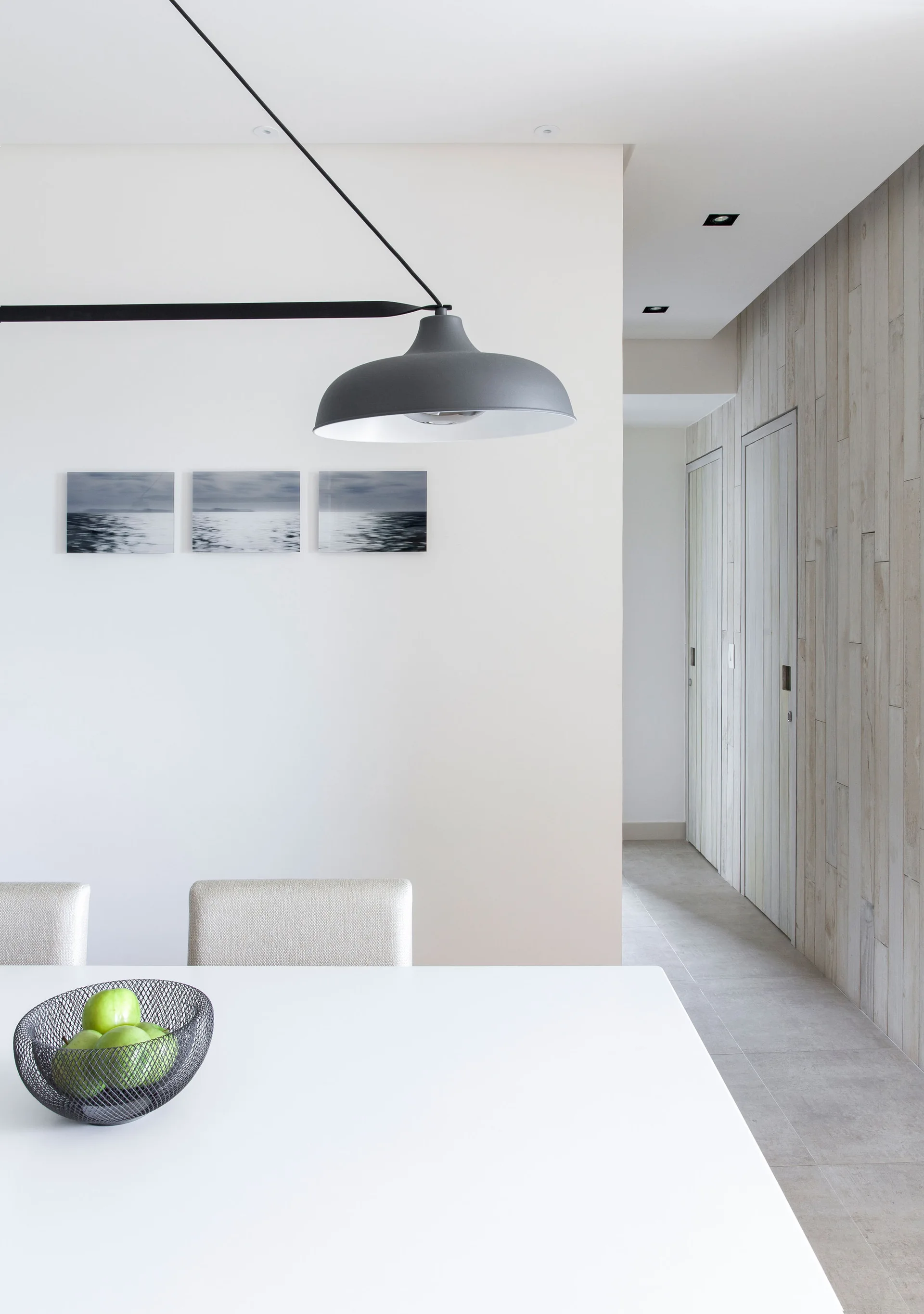 A minimalist dining room features a white table with green apples, a large grey pendant light, and a vertical wood plank accent wall.