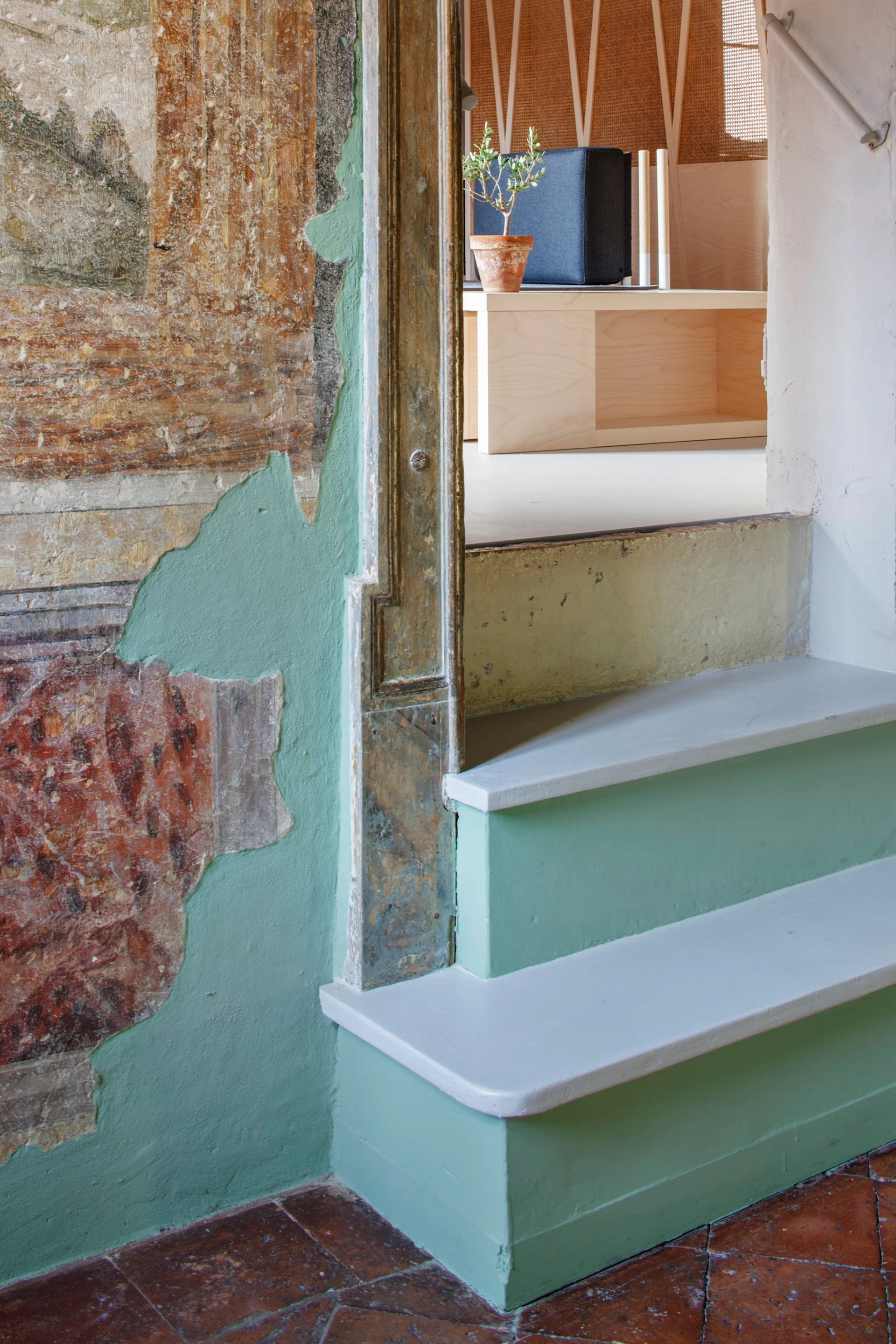 A painted sage green wooden staircase sits beside a distressed antique fresco wall with terracotta floor tiles visible below.