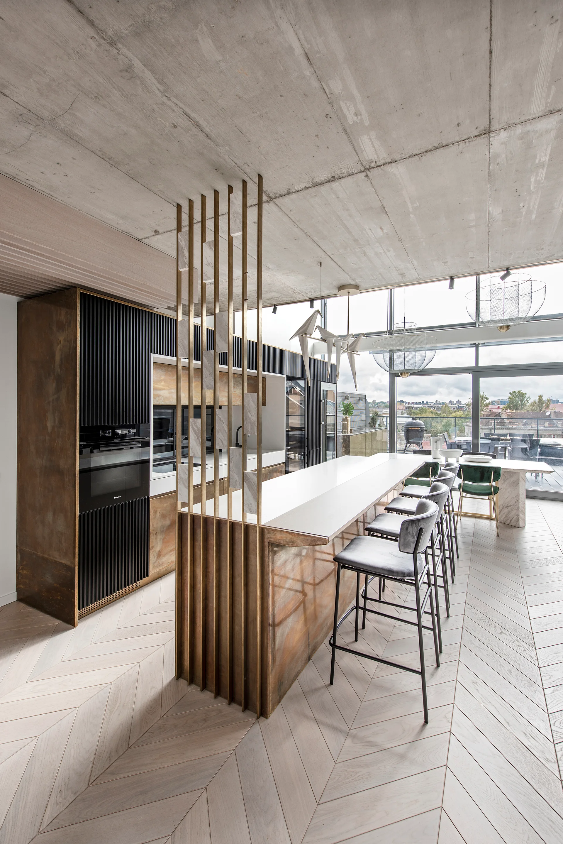 A modern open-plan kitchen with a metallic bronze island, vertical brass divider rods, light herringbone wood floors, and an exposed concrete ceiling.