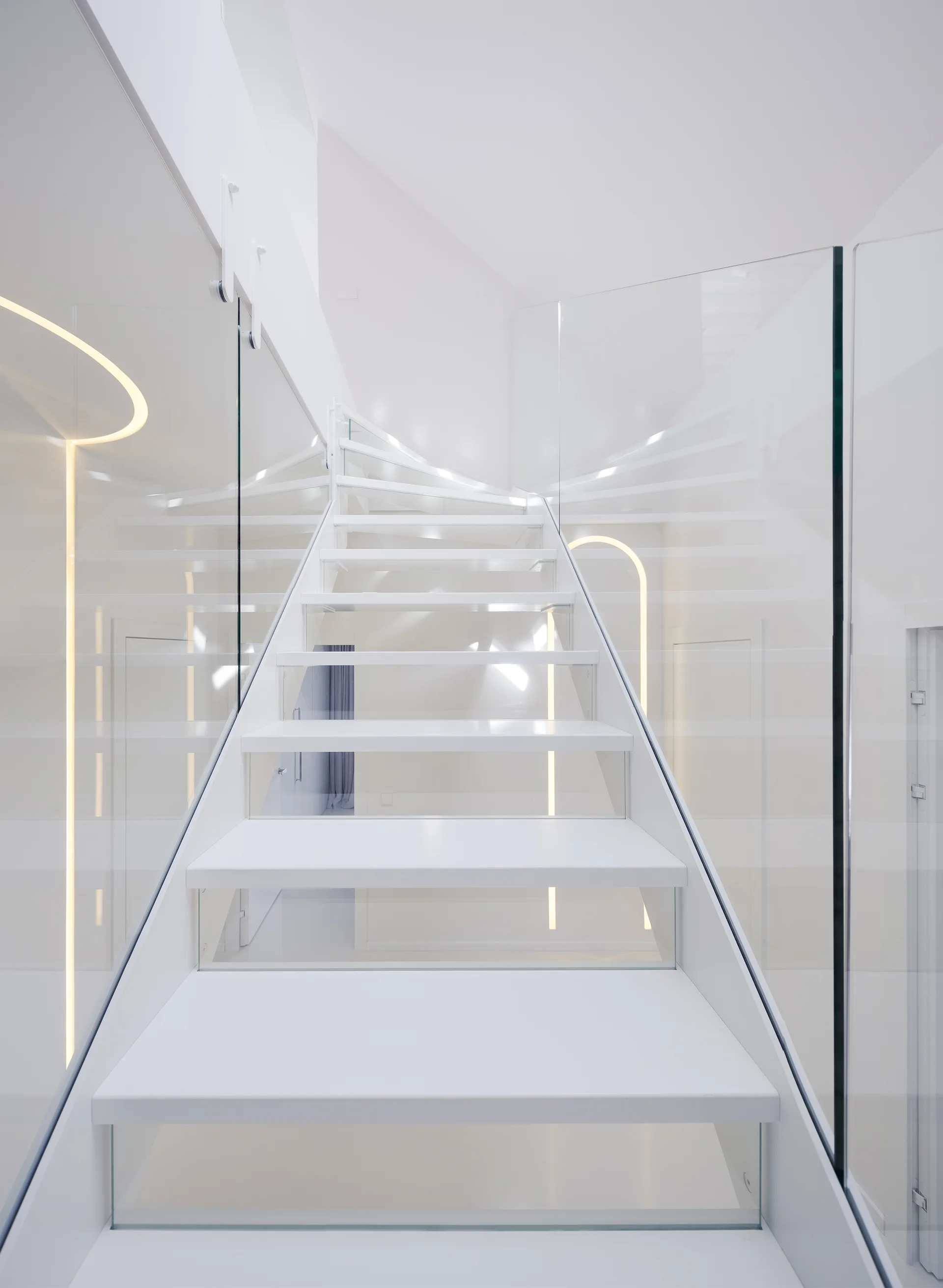 An all-white modern staircase with open treads, transparent glass panels acting as railings, and curved recessed LED strip lights on the white walls.