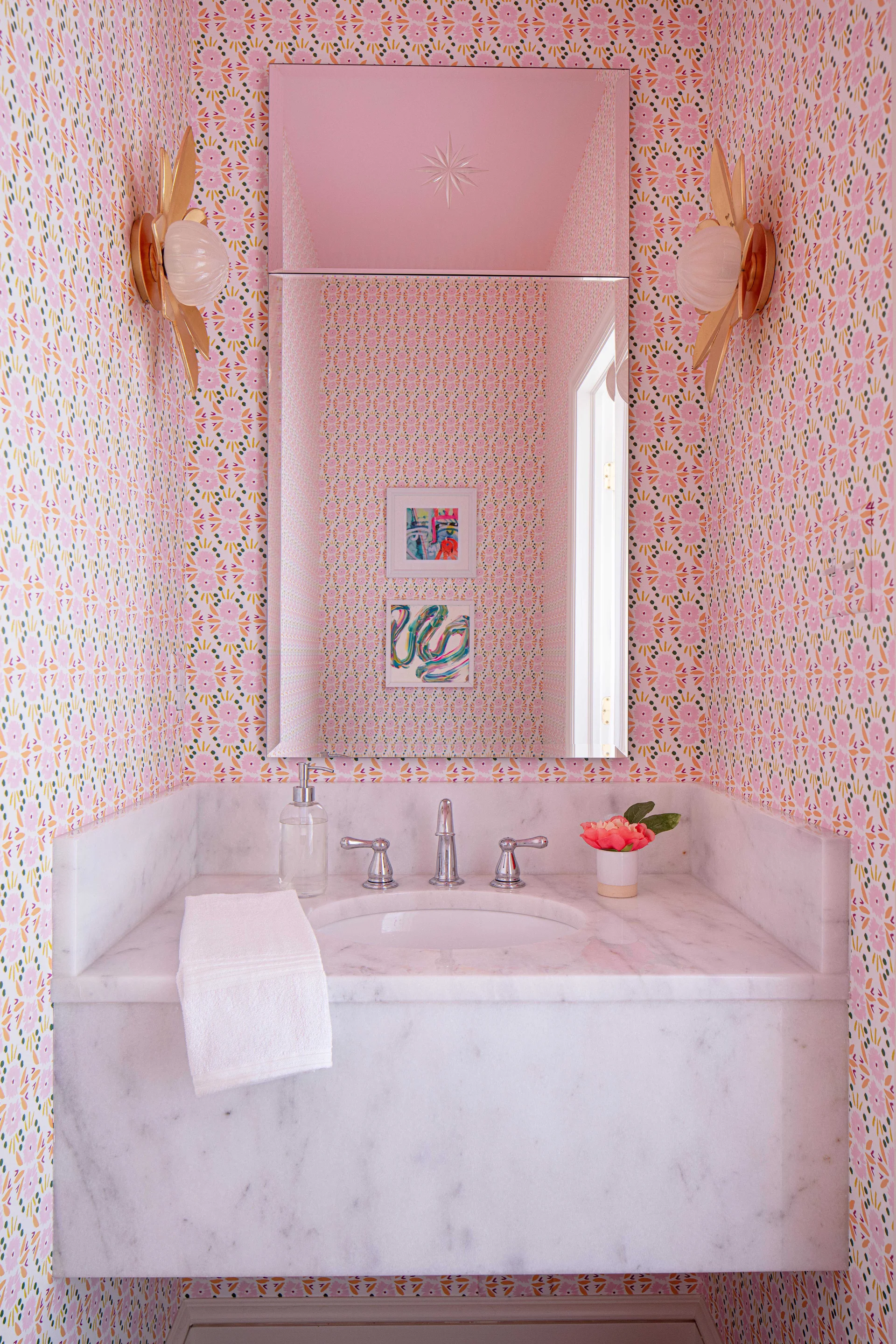 A powder room features pink floral wallpaper, a marble floating vanity, a tall rectangular mirror, and flower-shaped brass wall sconces.