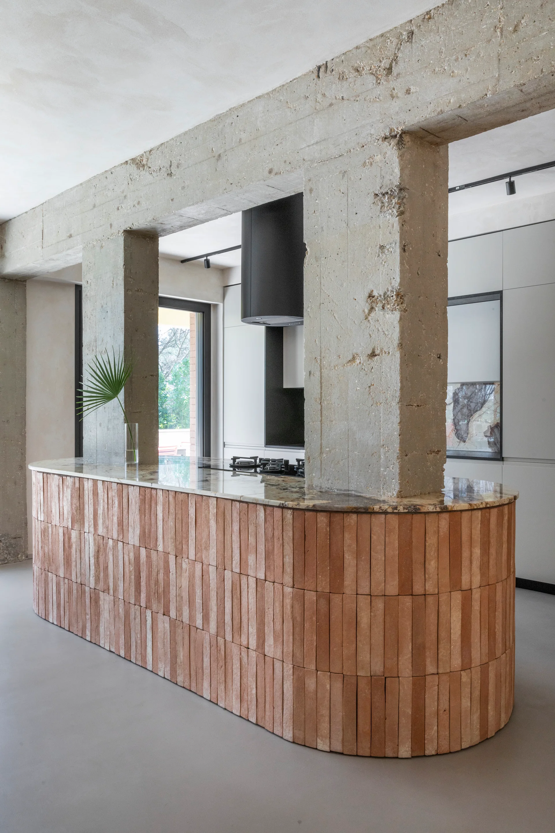 A curved kitchen island clad in vertical terracotta tiles stands beneath a raw concrete support column and beam next to white cabinetry.
