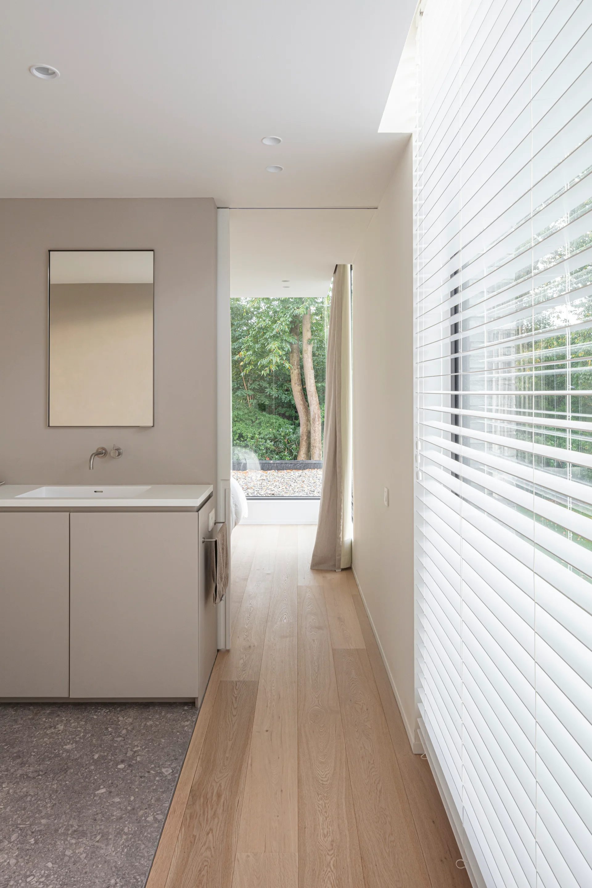 A minimalist taupe bathroom vanity with a mirror next to a light wood hallway with white horizontal blinds and a garden view.