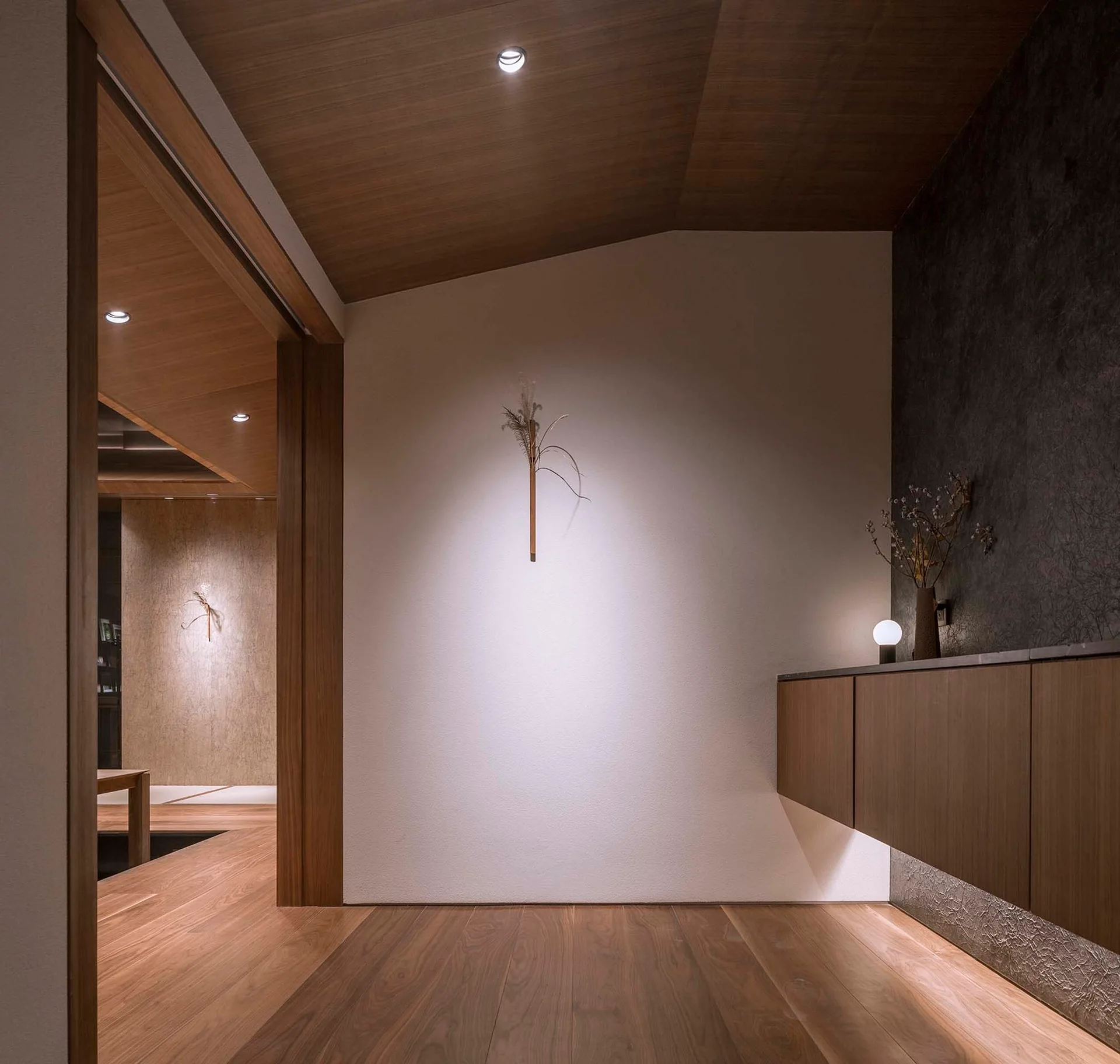 A minimalist entryway features a floating wooden cabinet against a dark textured wall, wood-paneled ceiling, and warm recessed spotlights.