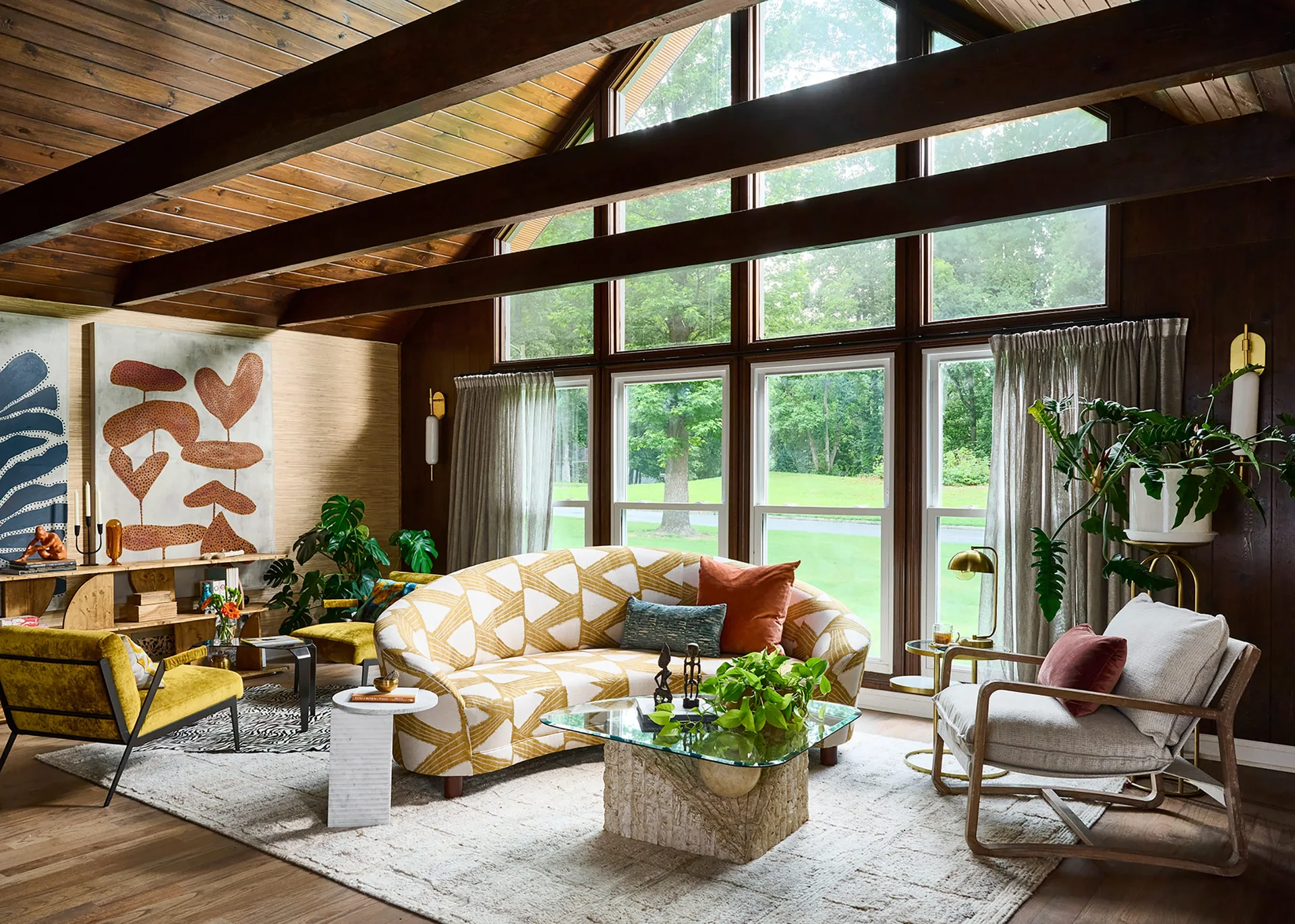 A living room with a vaulted wood ceiling and dark beams features a curved patterned sofa, mid-century chairs, and large windows looking out to trees.