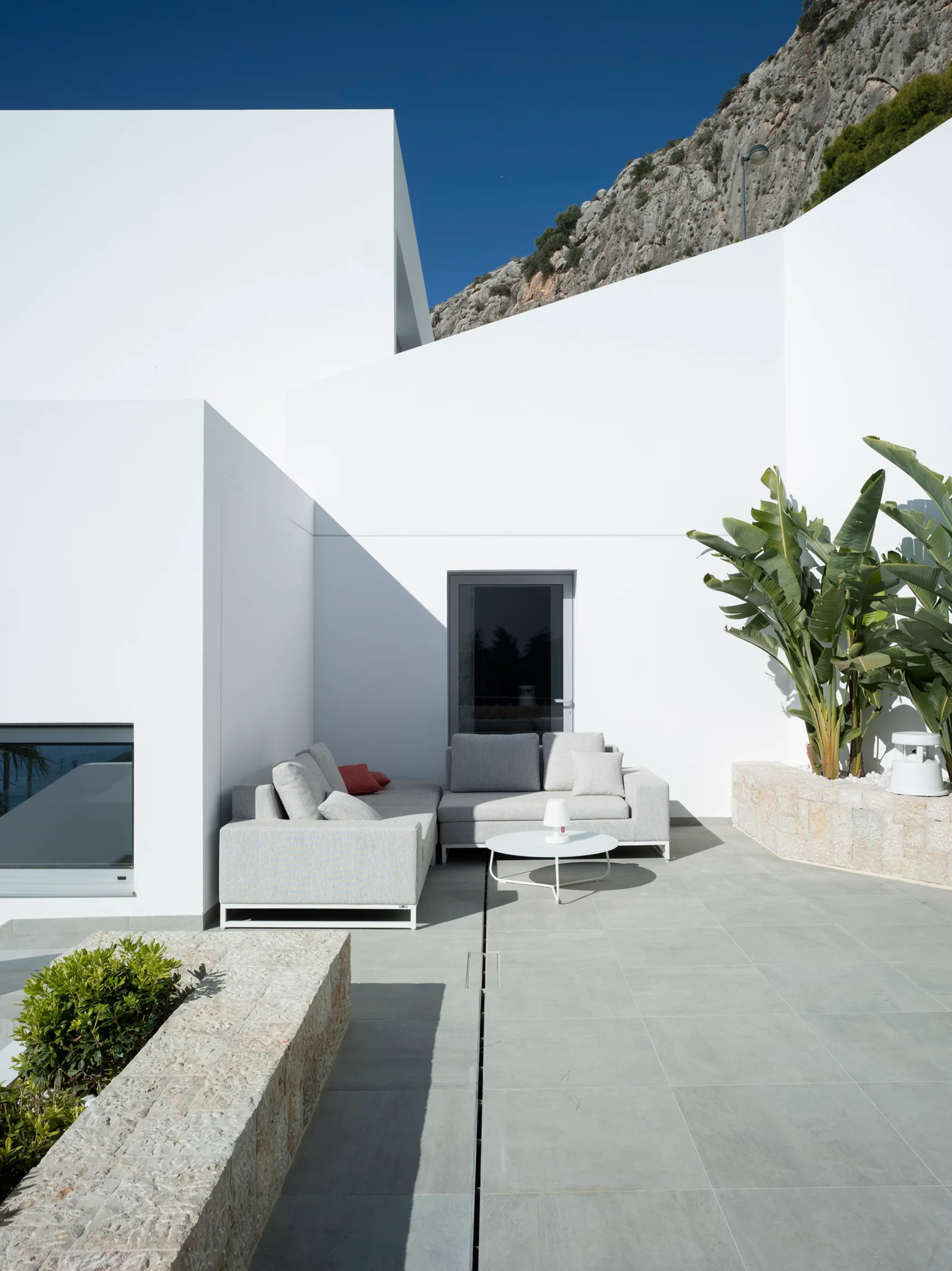 A light grey modular sectional sofa sits on a tiled patio next to built-in stone planters with leafy green plants against geometric white walls.